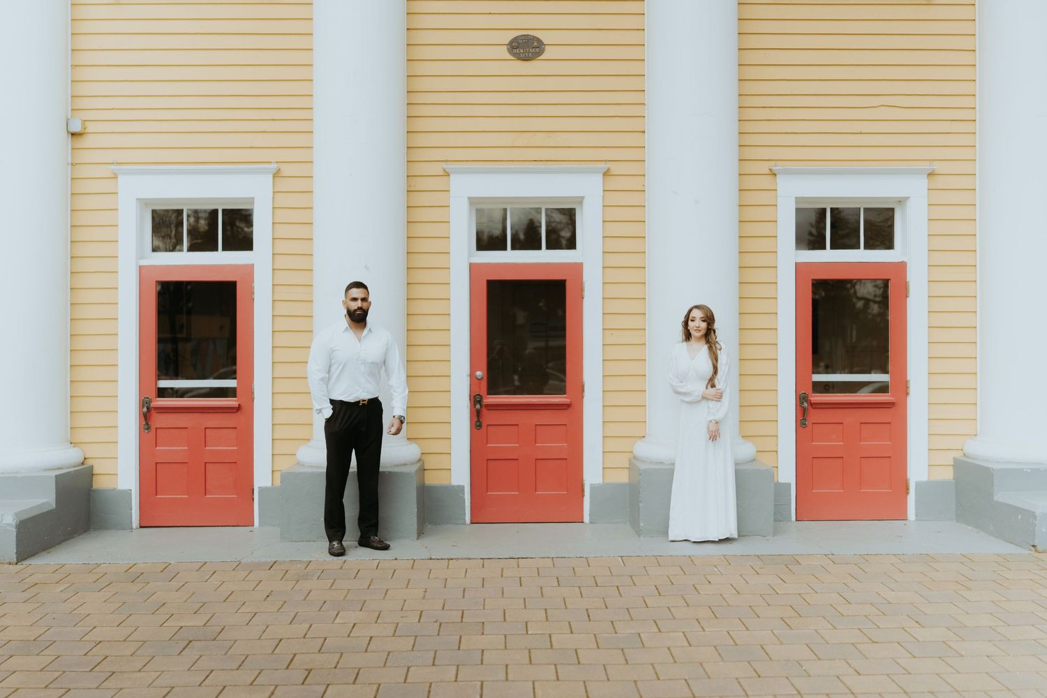 A minimalist wedding photo showing a couple standing between red doors of a yellow colonial building.