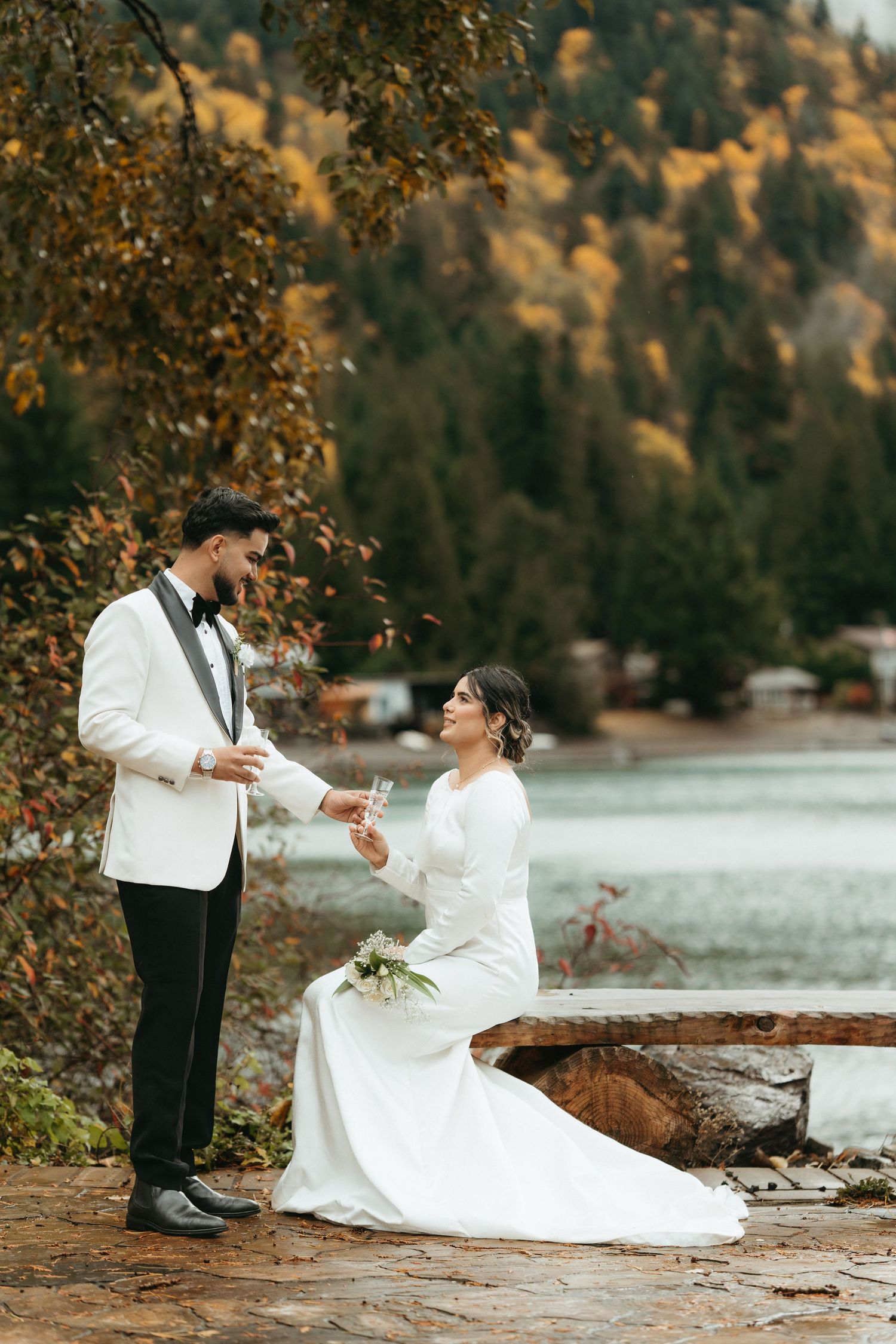A couple in wedding attire share a romantic moment by a serene lakeside surrounded by autumn foliage.