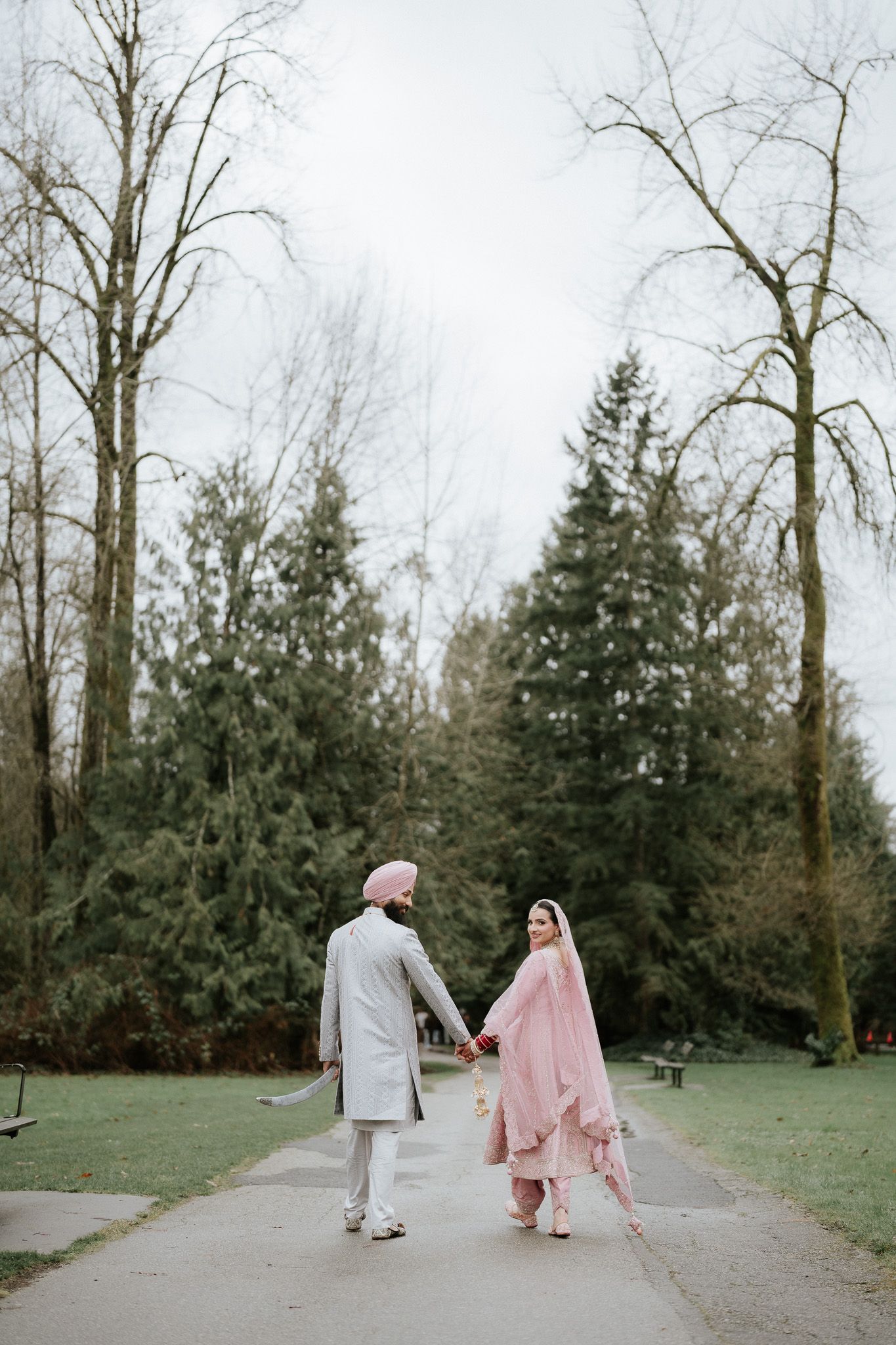A couple walks hand in hand down a peaceful path surrounded by evergreen trees and bare branches in winter.