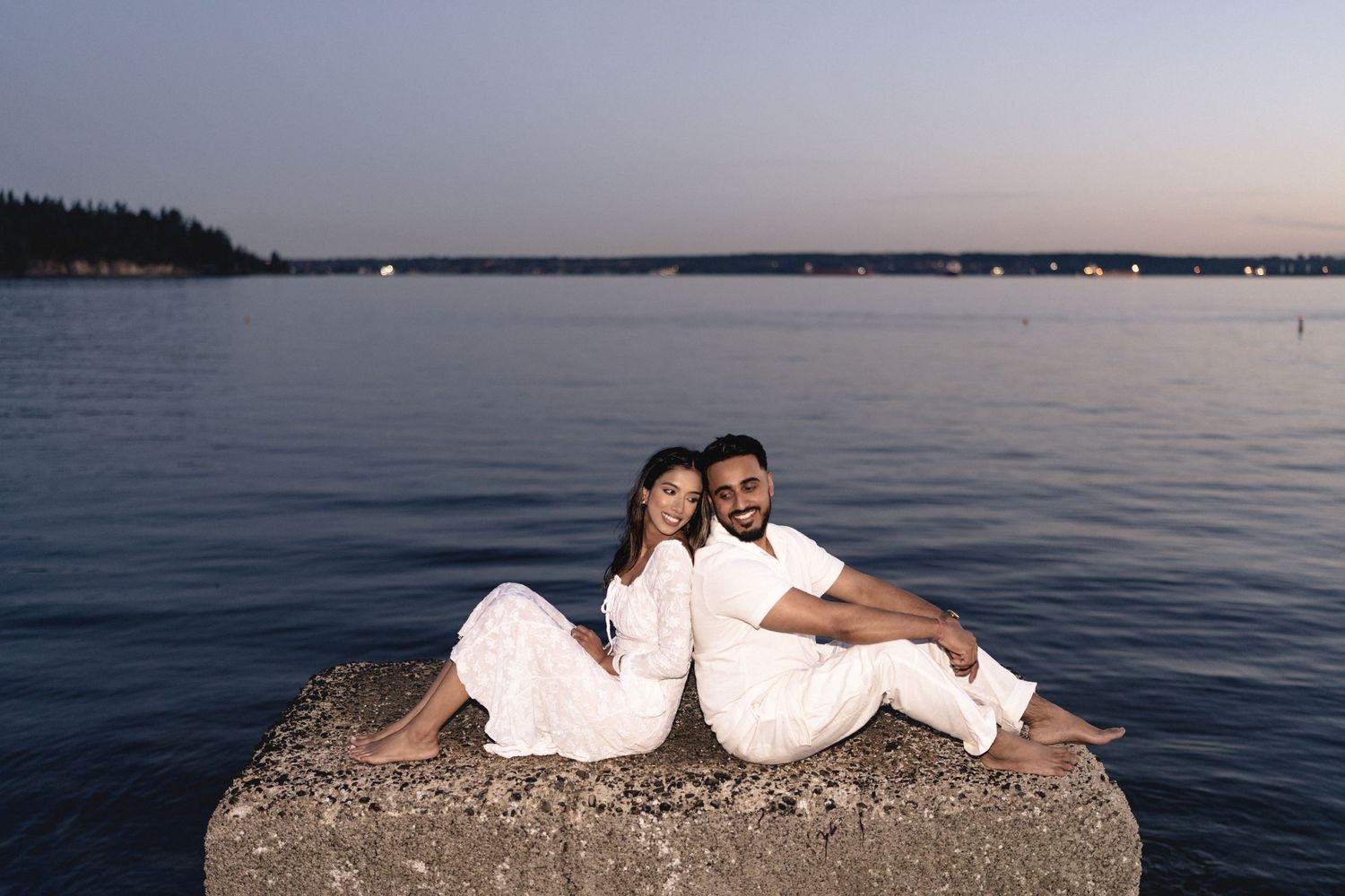 A couple in white clothing sits together on a rocky shore at sunset by a calm lake.