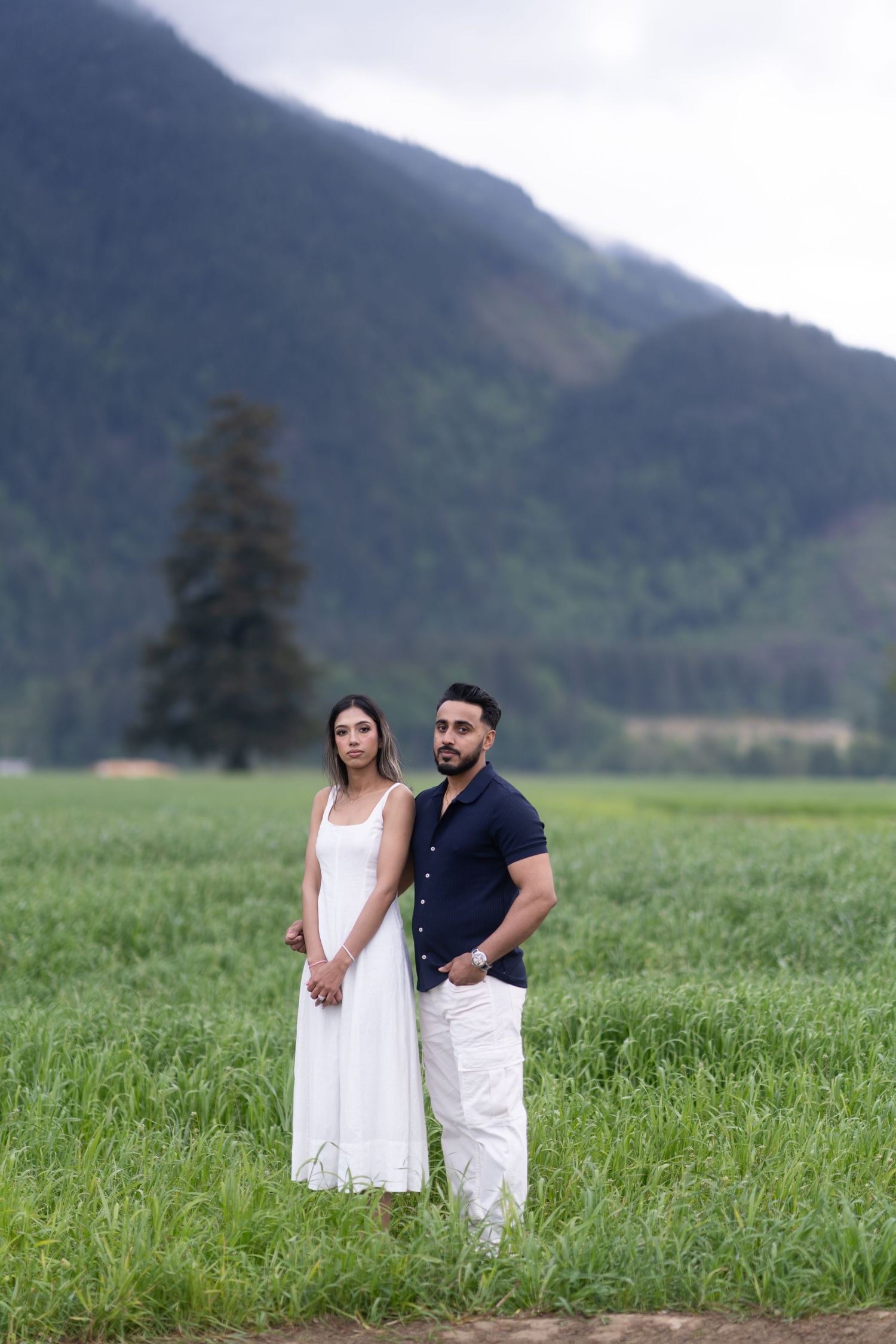 A couple stands together in a lush green field with misty mountains in the background.