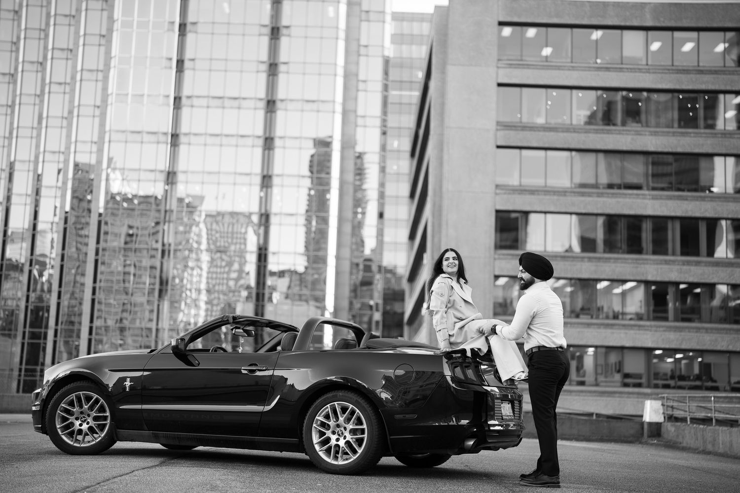 A sleek black luxury car is parked in front of modern glass buildings in a monochrome urban setting.