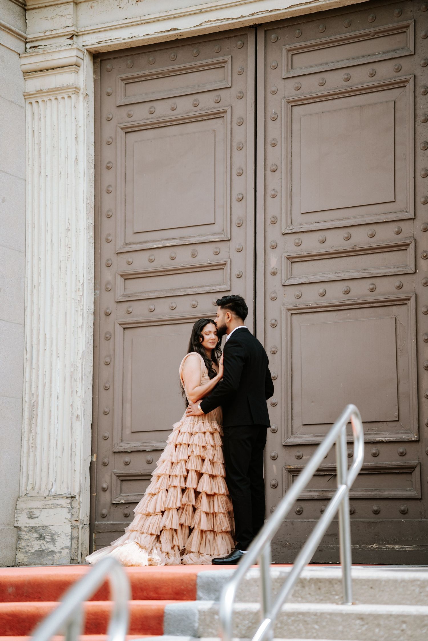 Couple in formal wear share intimate moment by ornate building entrance.