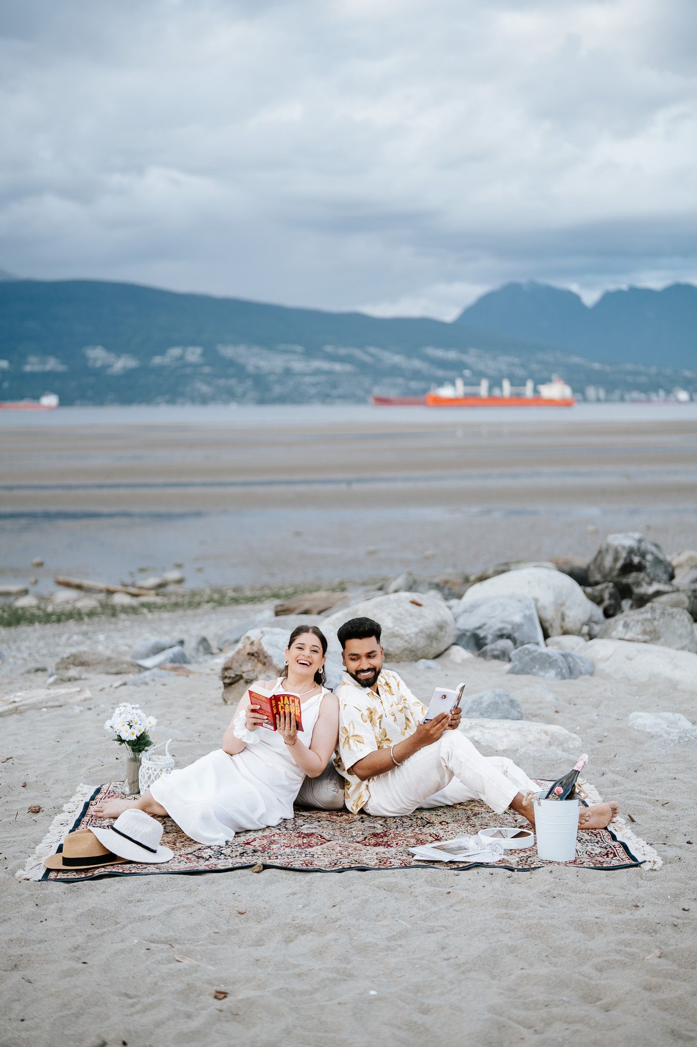 Couple in white outfits enjoying a beach picnic on a rug with books and drinks, mountains in background.