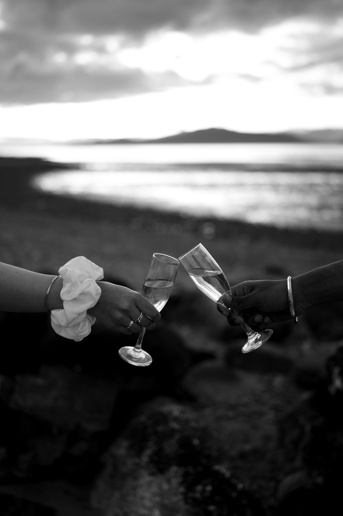 Two hands clinking champagne glasses at a beach sunset in black and white, celebrating a romantic occasion.