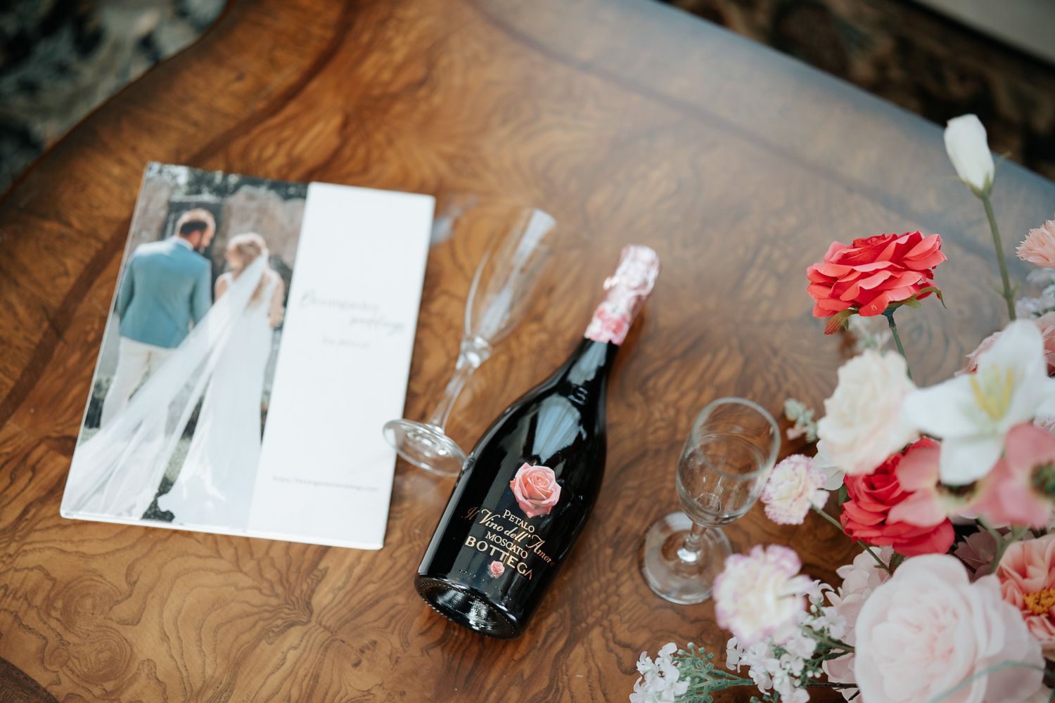 Elegant wedding flat lay with champagne bottle, floral arrangement, glasses, and couple photo on wooden table.