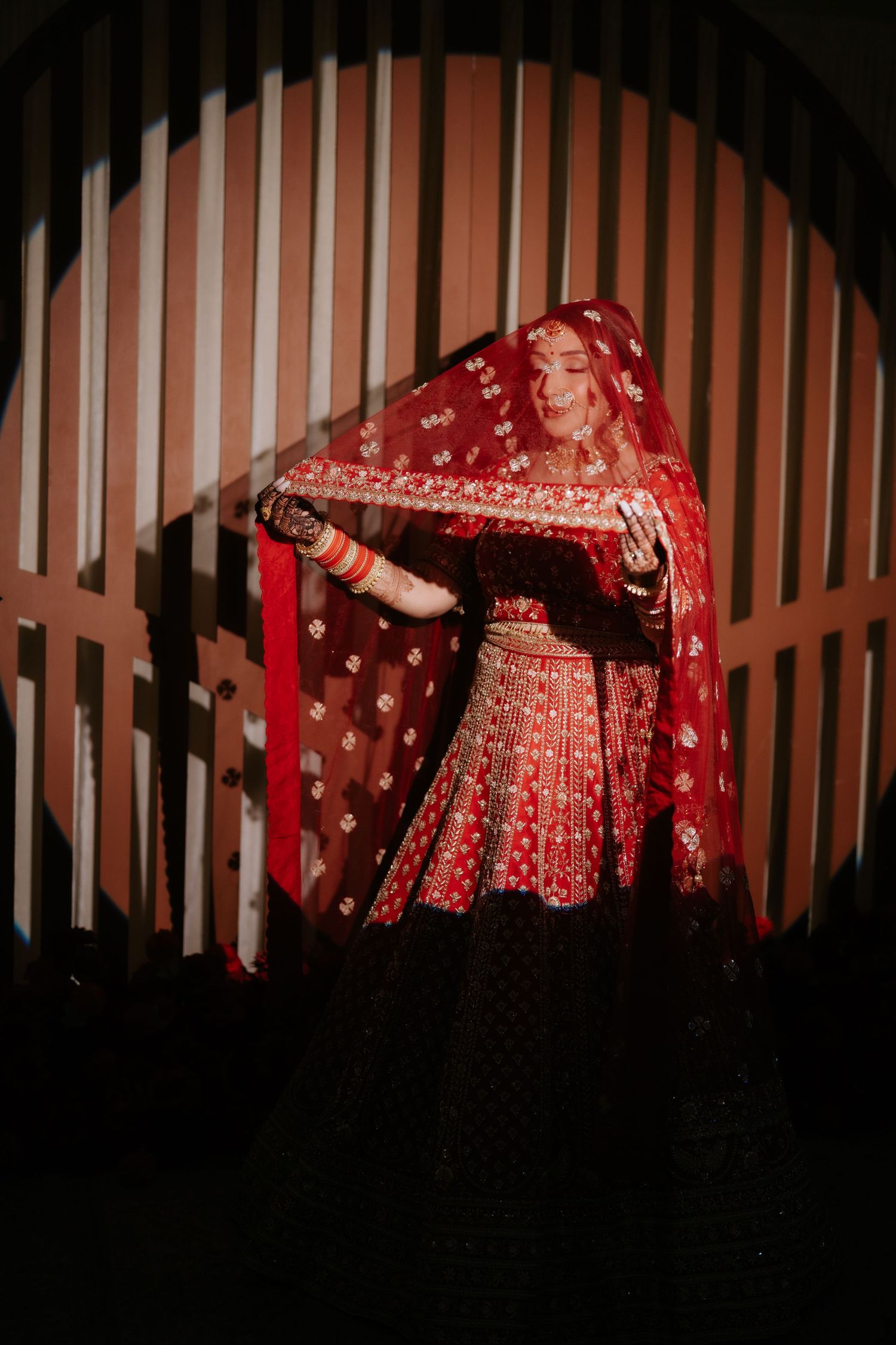 A bride in a stunning red embroidered lehenga and dupatta poses gracefully in a dimly lit arched doorway.