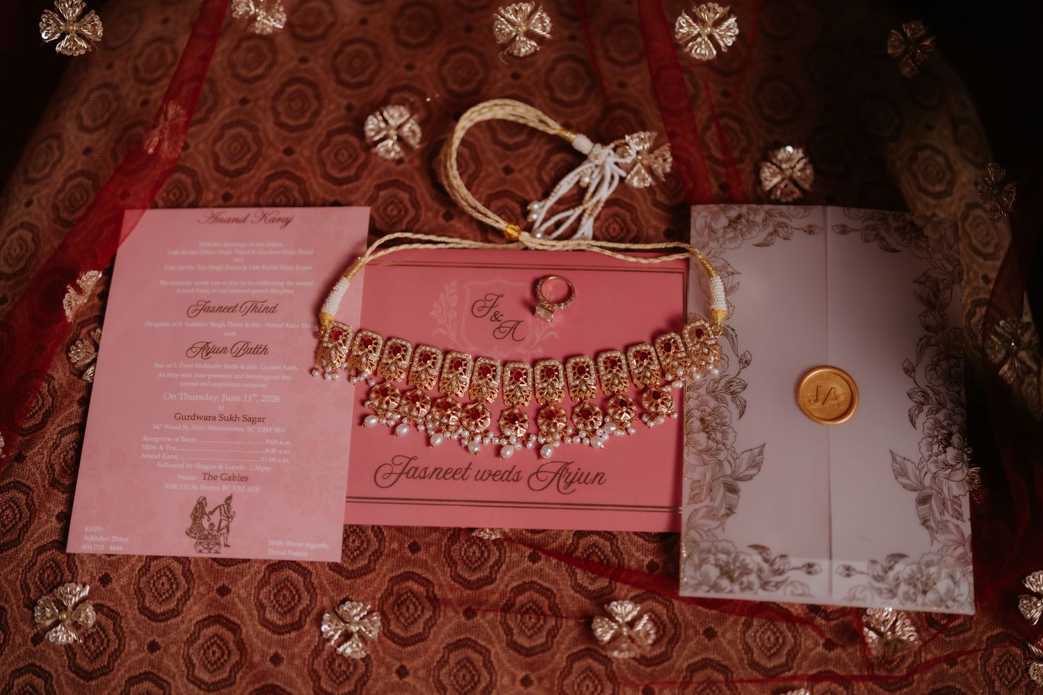 Pink Indian wedding invitation set with gold necklace jewelry on red fabric backdrop.