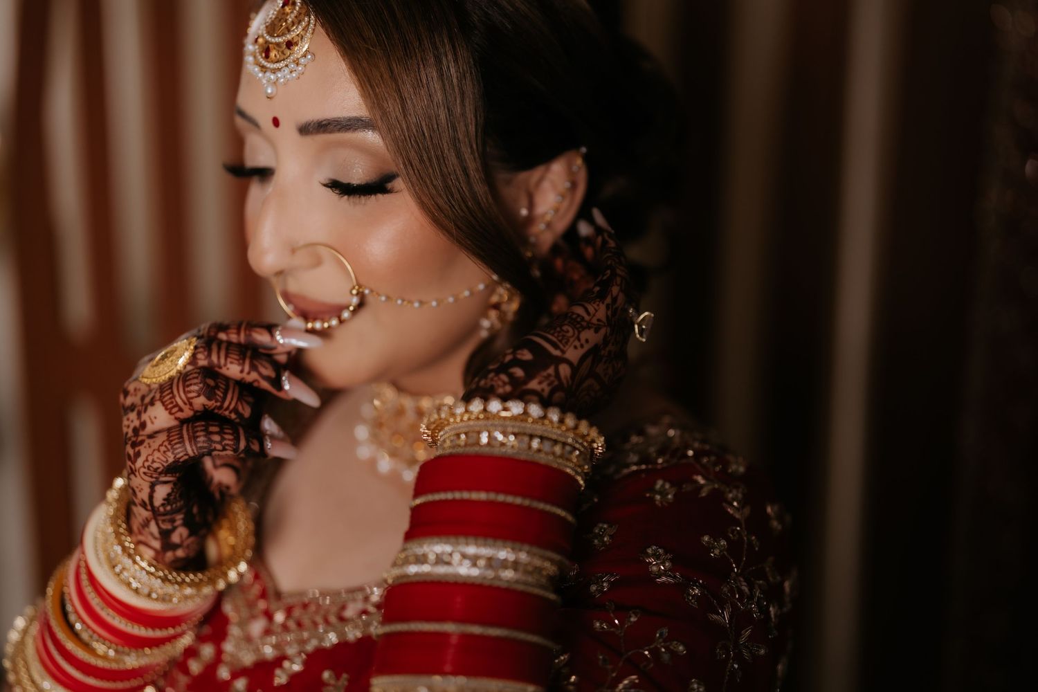 Indian bride in red lehenga adjusting nose ring with mehndi hands and gold jewelry.