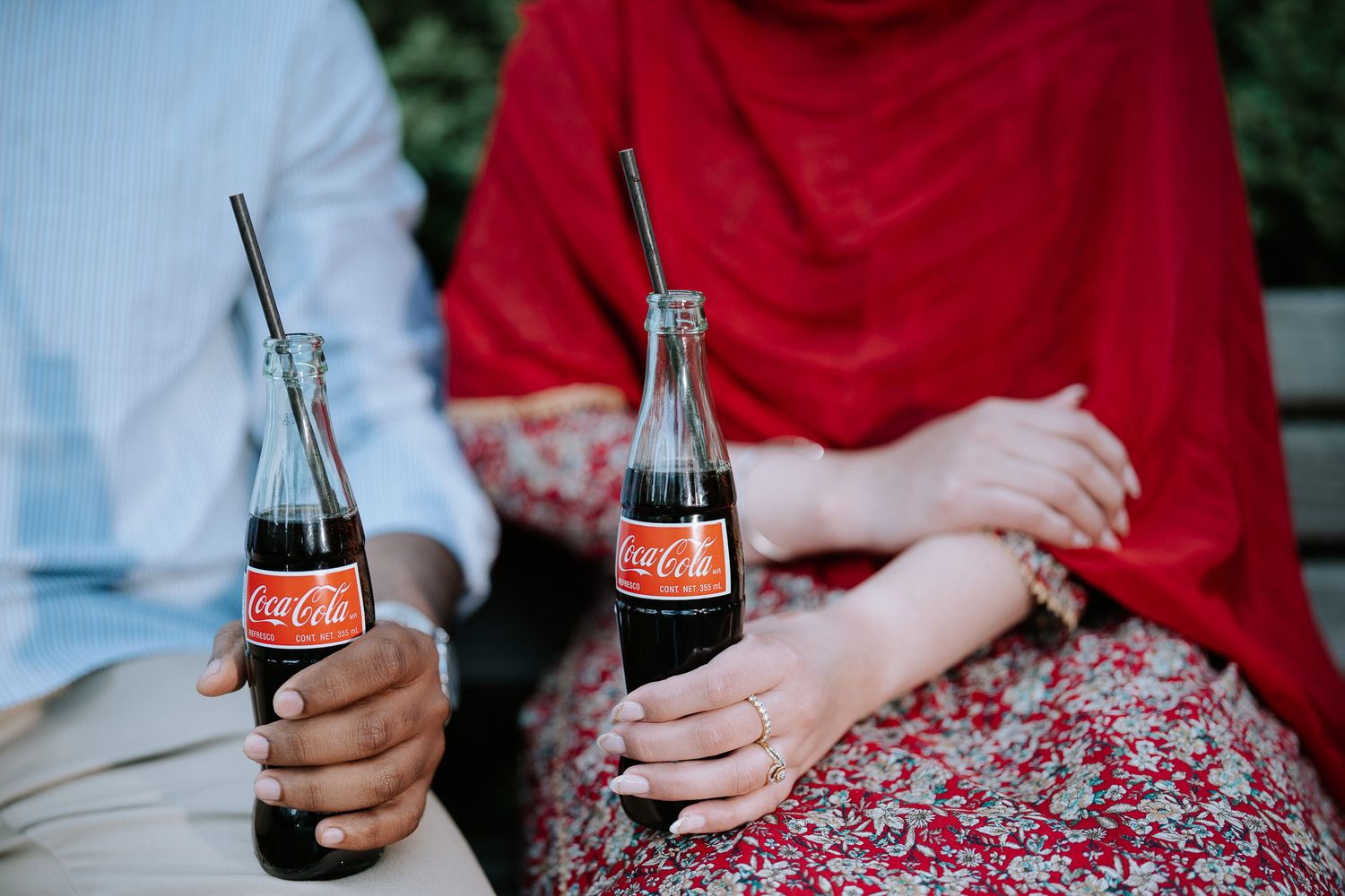 Couple sharing Coca-Cola bottles with metal straws outdoors, woman in red hijab and floral skirt.