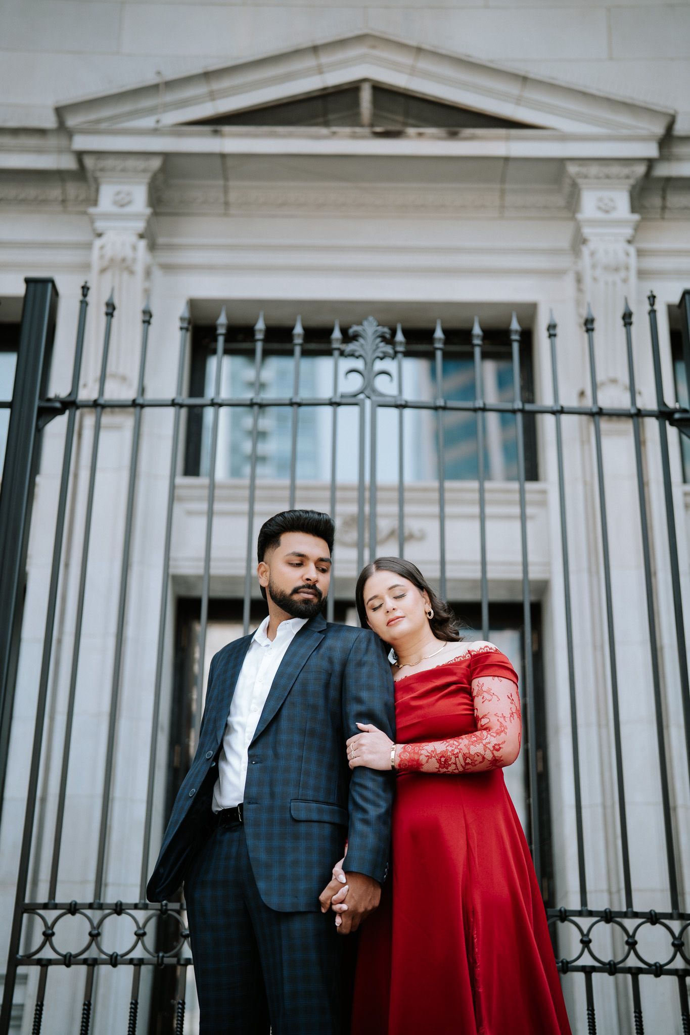 Elegant couple posing before ornate building, man in suit and woman in red off-shoulder gown.