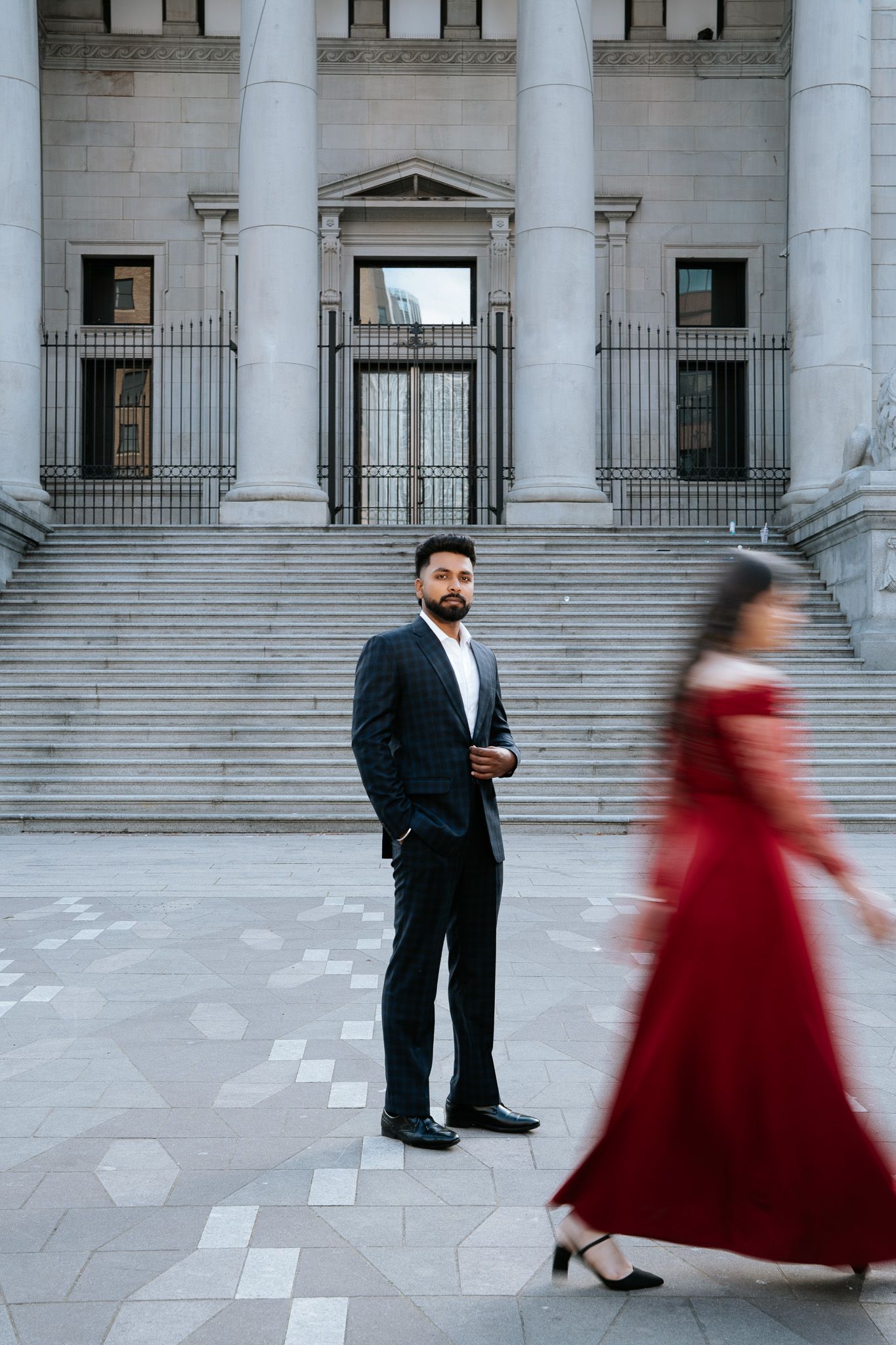 Man in dark suit stands before grand building steps as woman in red gown walks past.