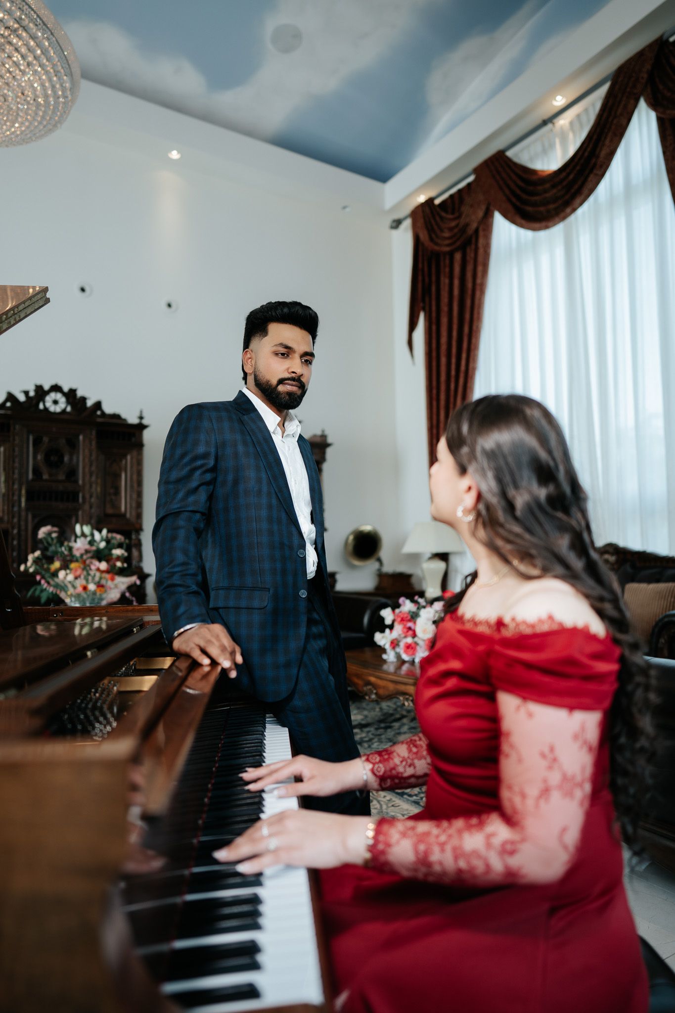 Bearded man in blue suit watches woman in red lace dress playing an upright piano in elegant room.