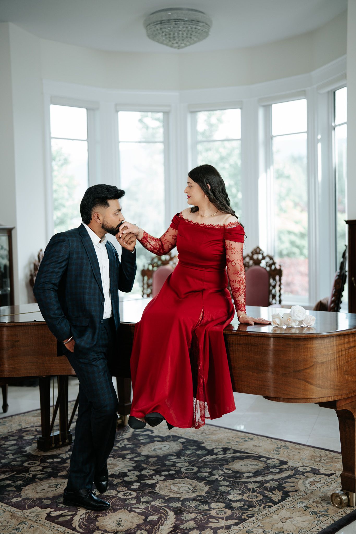 Man in dark suit kisses woman's hand as she sits on grand piano in elegant room.