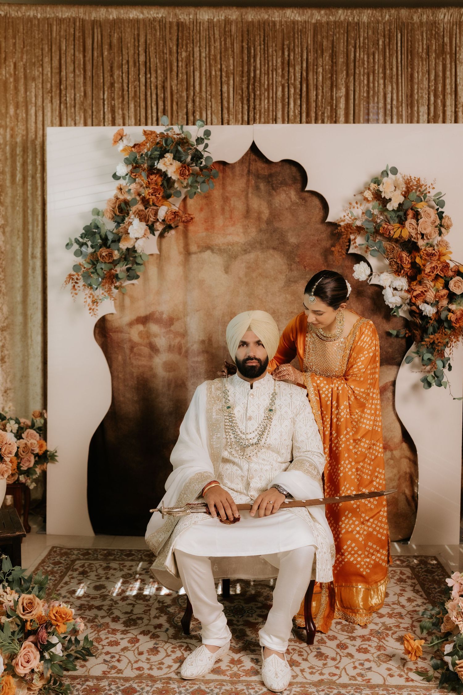 Sikh couple in traditional wedding attire pose before ornate floral backdrop, groom seated in white sherwani, bride in orange lehenga.