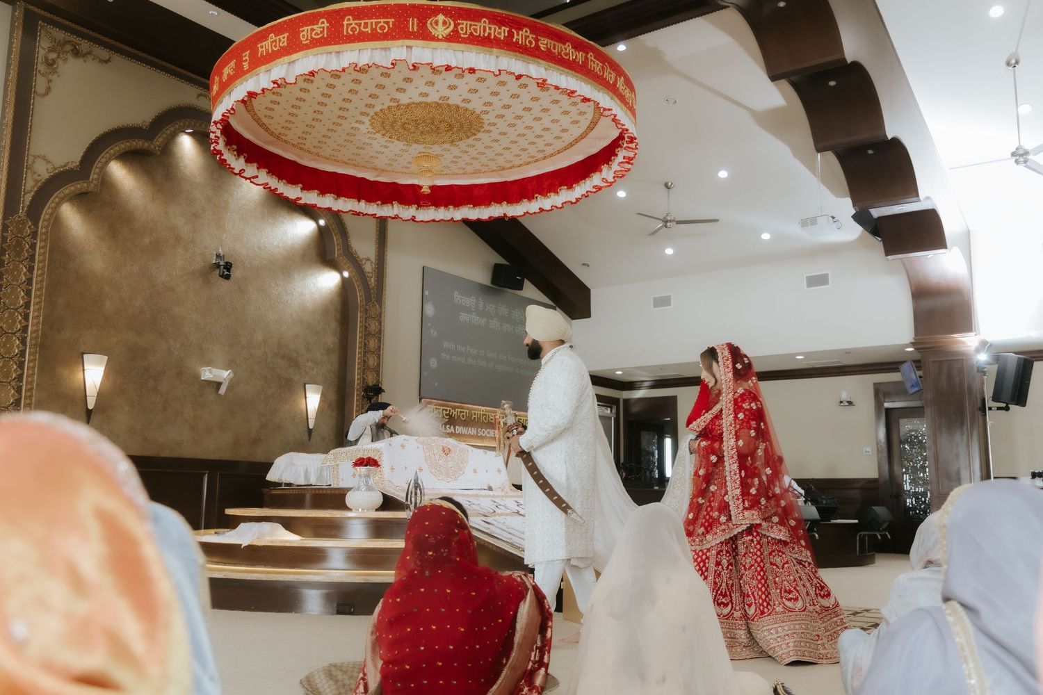Sikh wedding ceremony with bride in red lehenga and groom in white sherwani walking under decorative red canopy.
