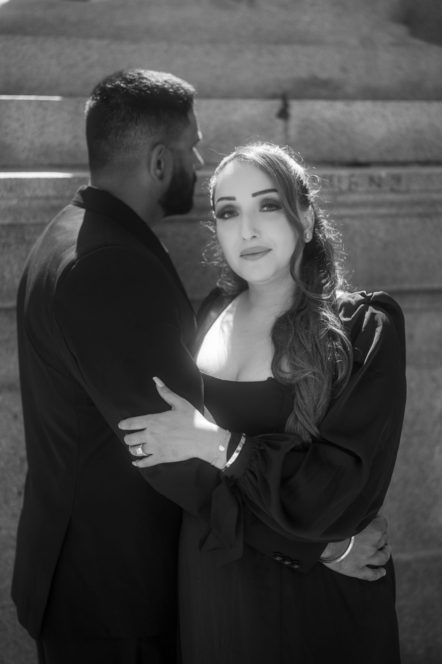 Black and white photo of a couple embracing outdoors, woman in black dress gazes at camera while man faces away.