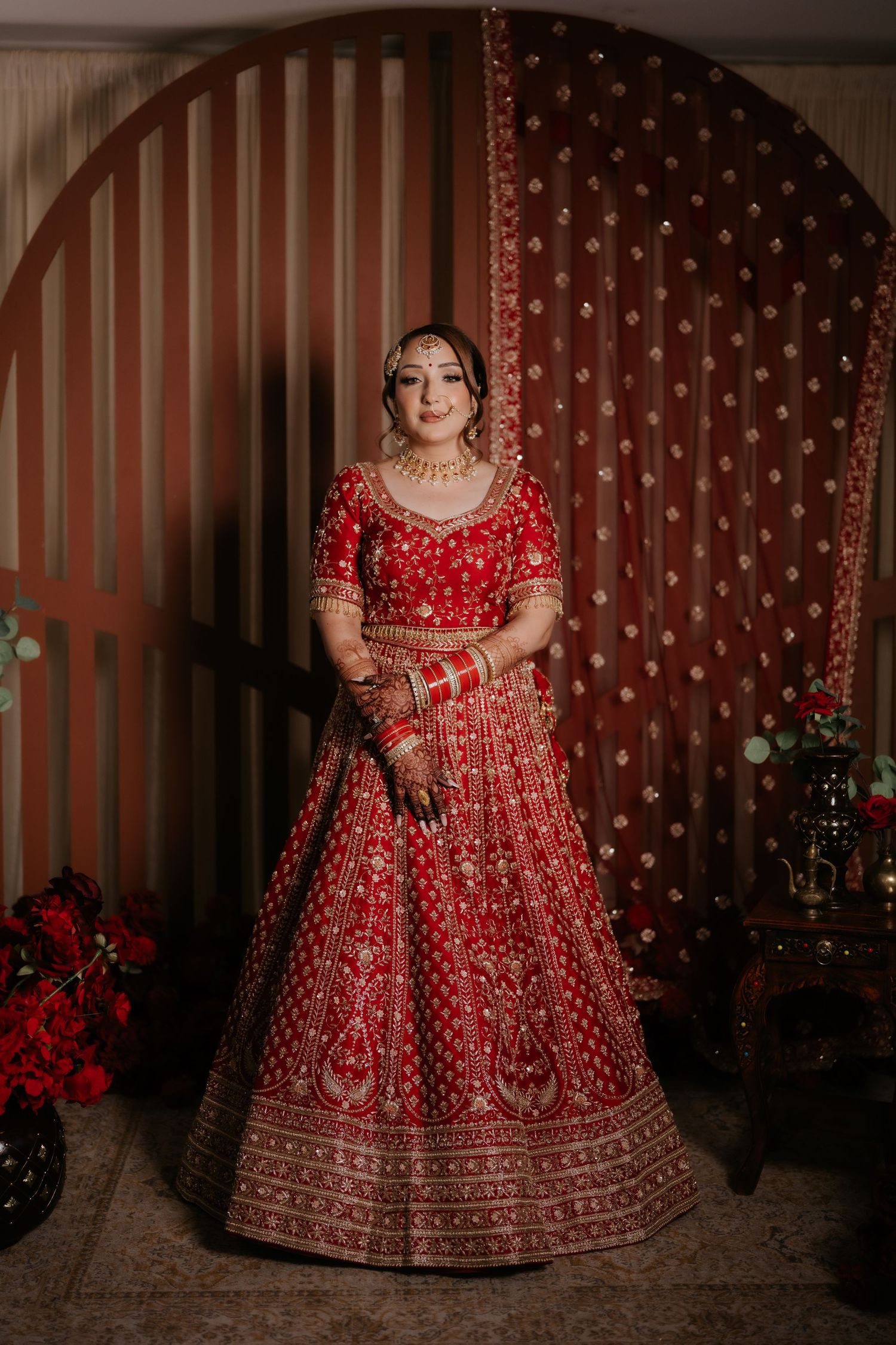 Bride in red embroidered lehenga with gold jewelry poses gracefully in front of decorative arch backdrop.