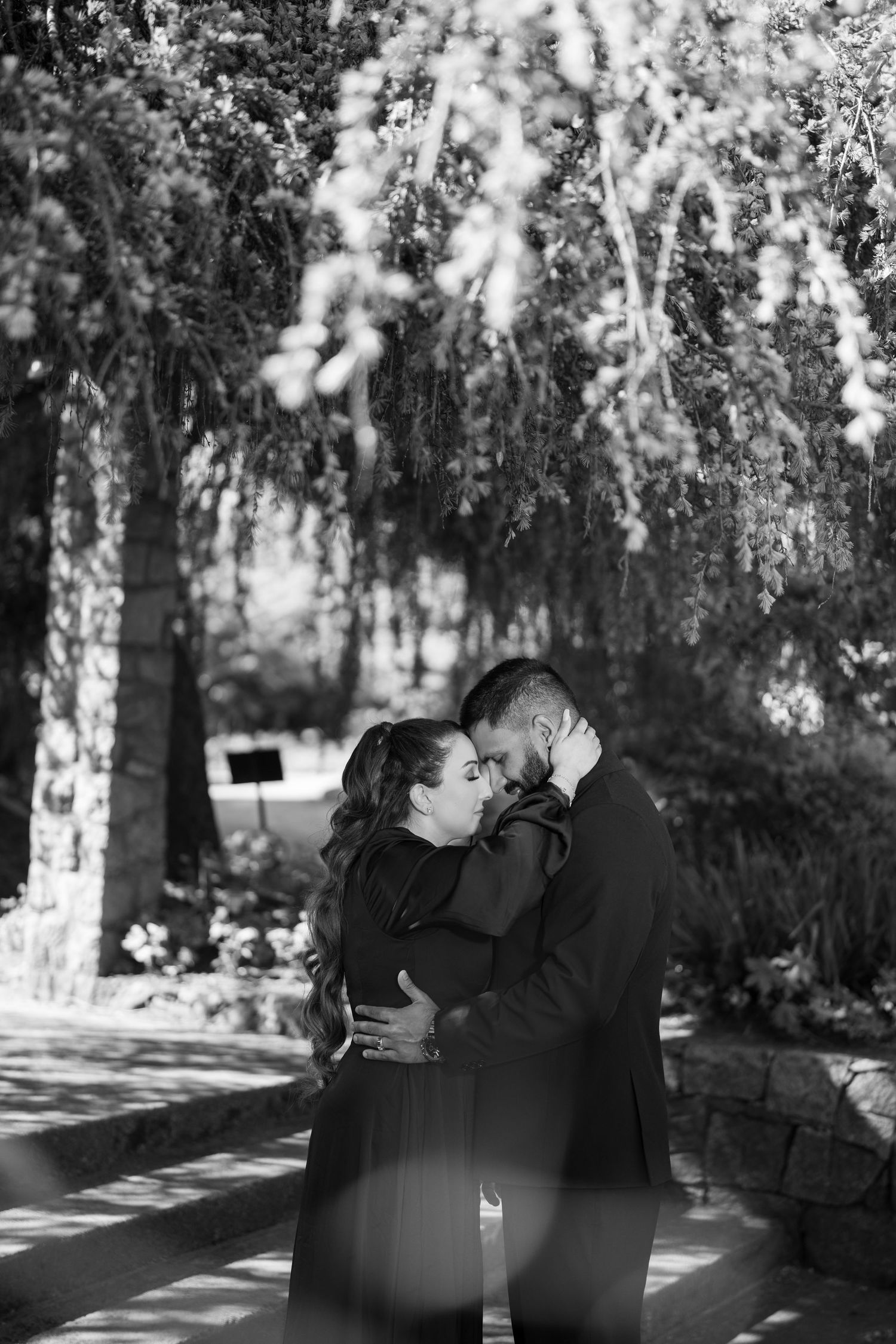 Black and white photo of a couple embracing and kissing outdoors under lush weeping trees in a garden setting.