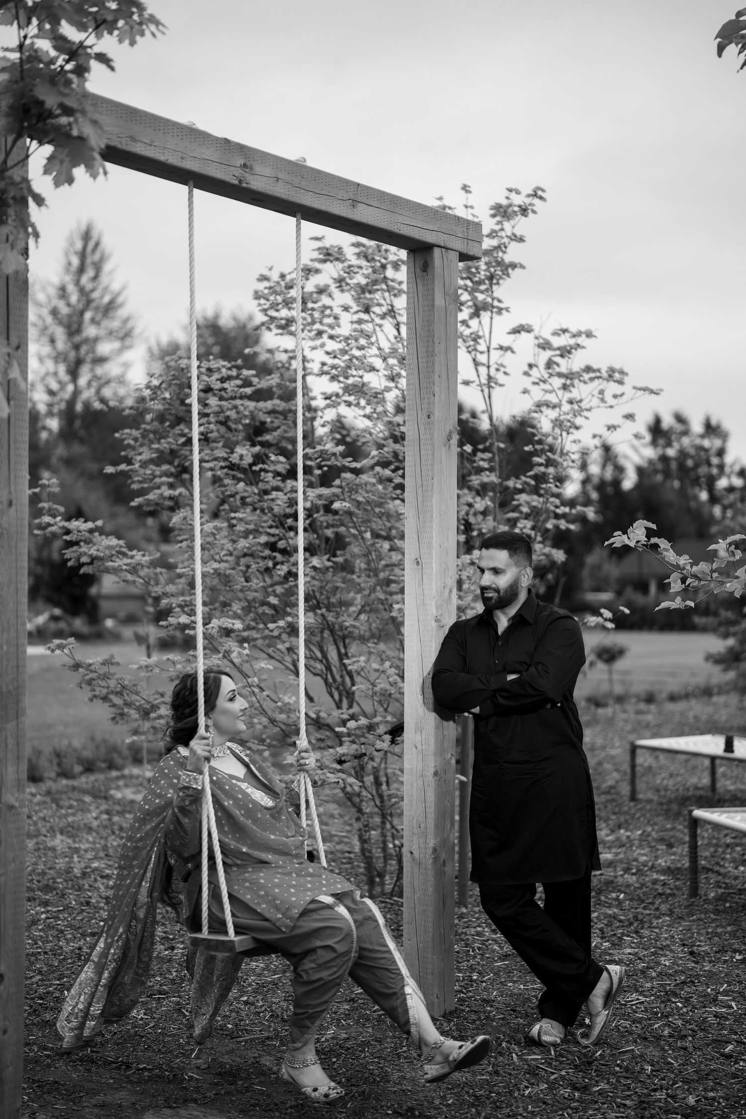 Black and white photo of a couple, woman sitting on wooden swing while man leans against frame in garden.
