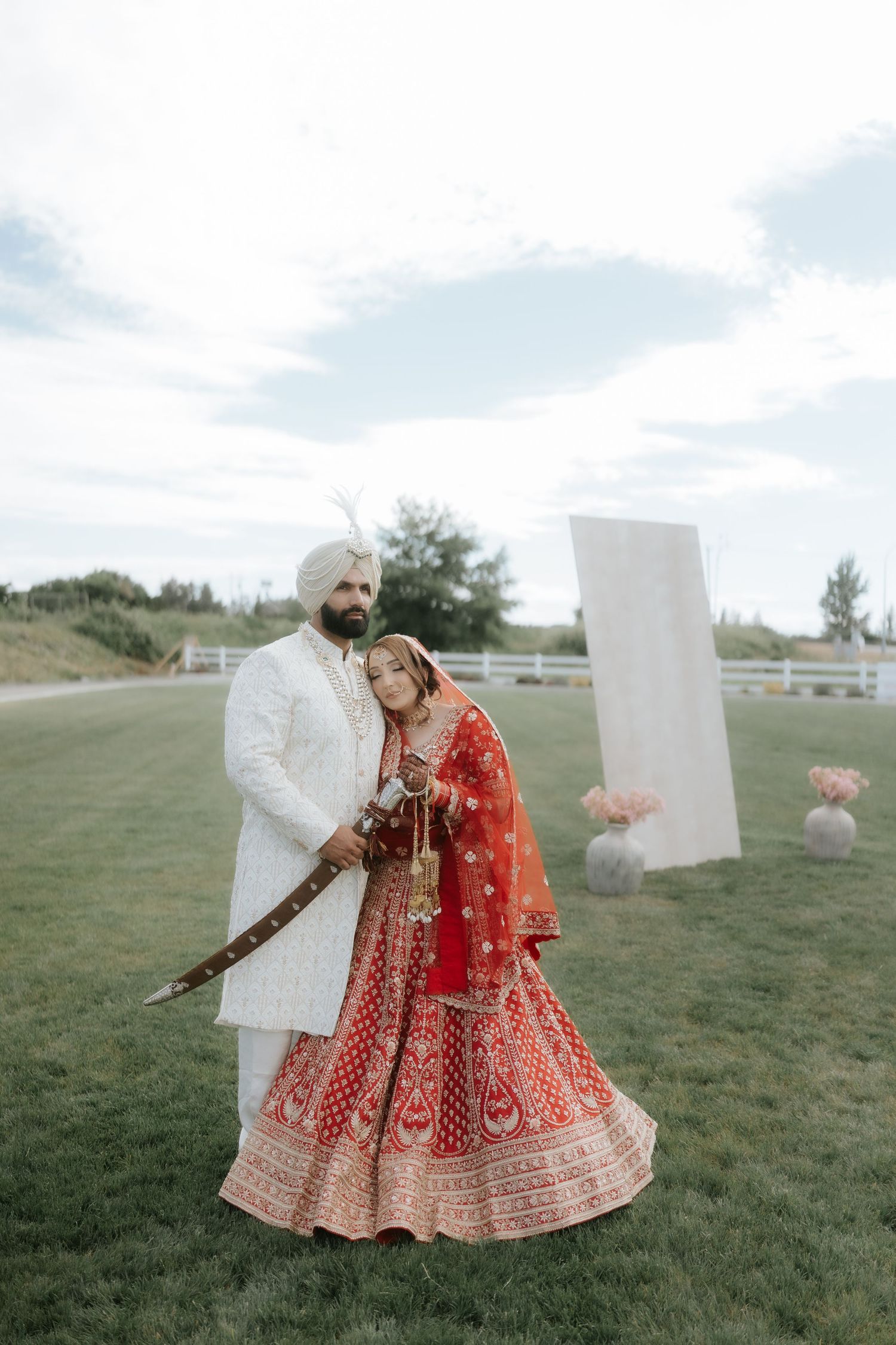 Sikh bride in red lehenga and groom in white sherwani and turban pose outdoors at traditional wedding ceremony.