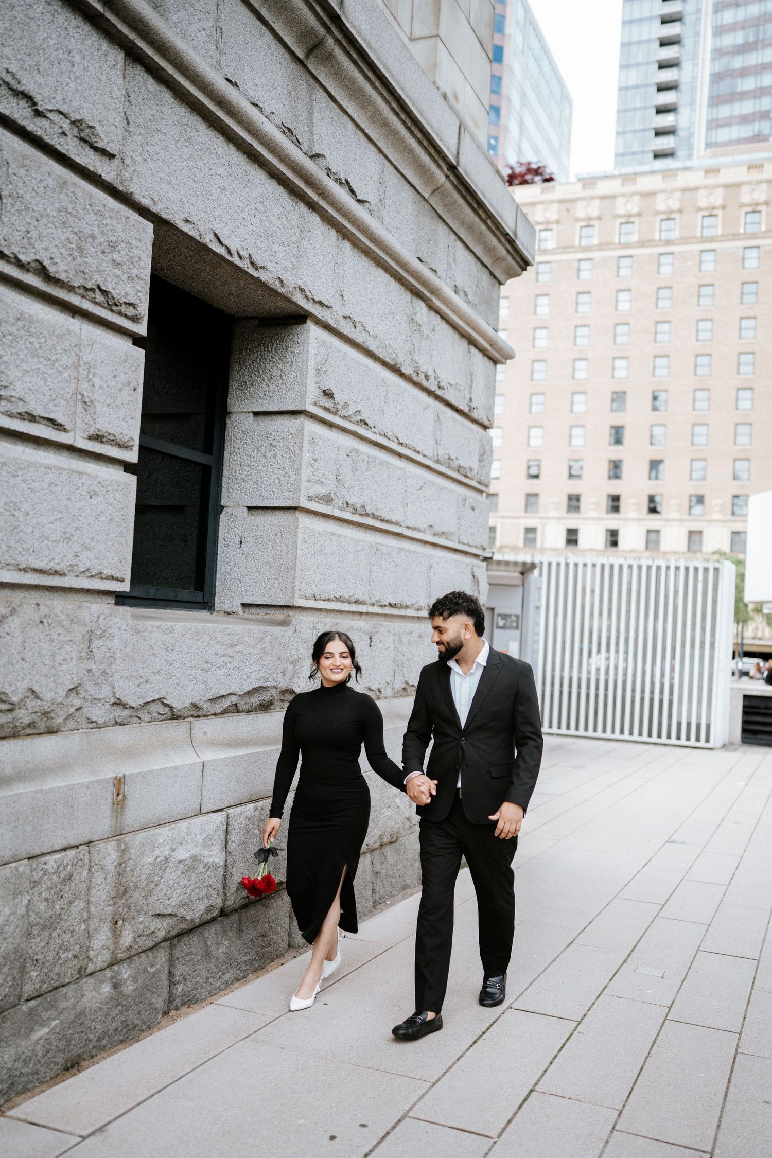 Couple in formal wear walks along stone building wall in urban setting.