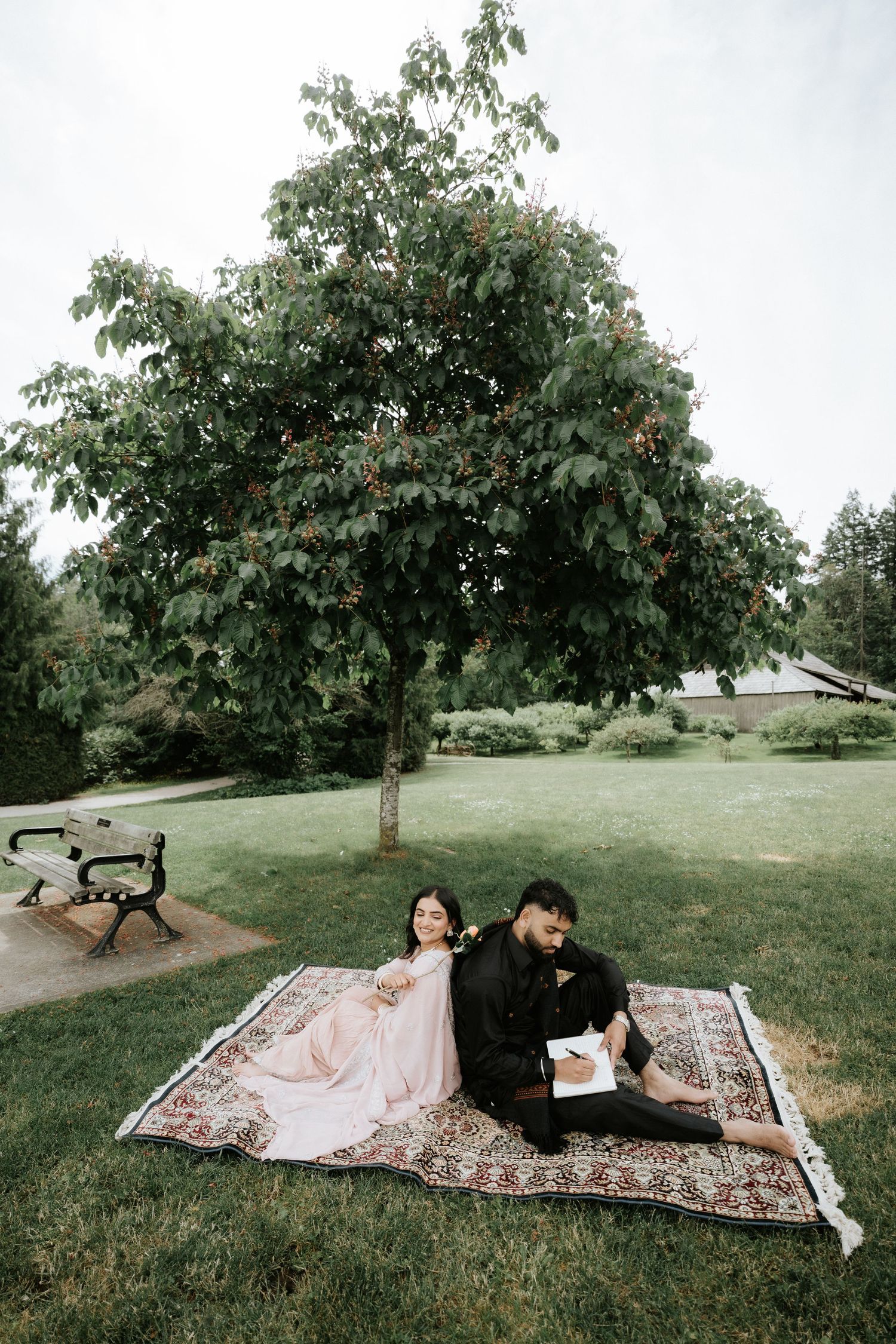 A couple relaxes on a patterned blanket under a leafy tree in a peaceful outdoor setting.