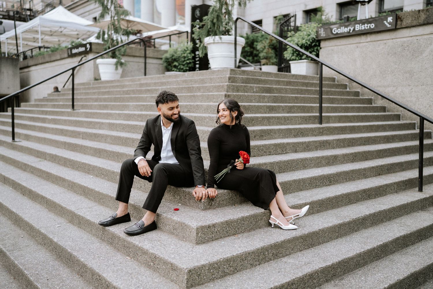 A couple sits casually on wide stone steps in formal black and white attire.