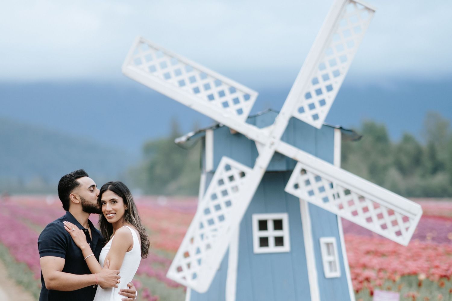 Couple poses romantically in front of decorative white windmills and pink flower fields during golden hour.