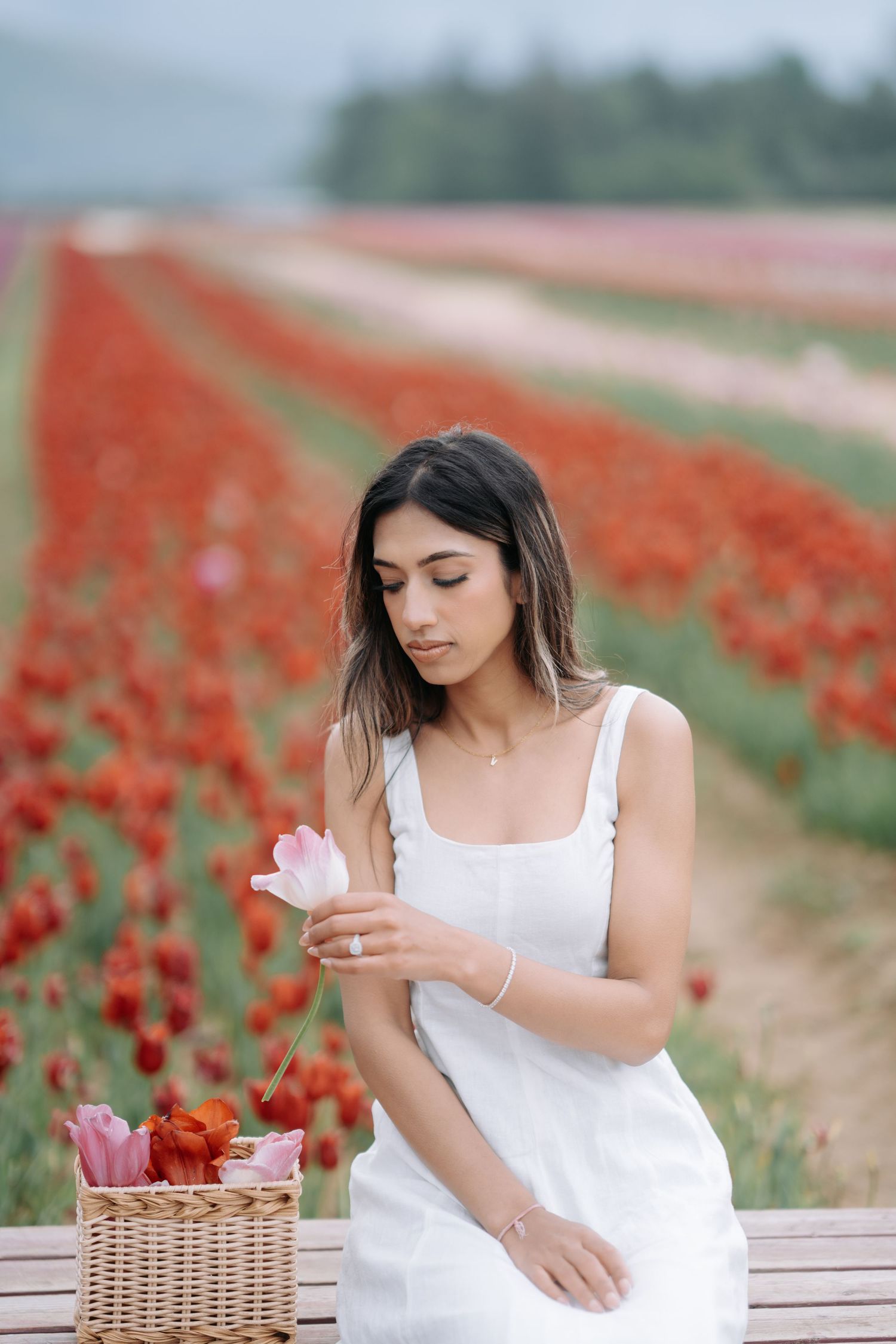 A woman in a white dress sits thoughtfully among vibrant red tulip fields on a sunny spring day.