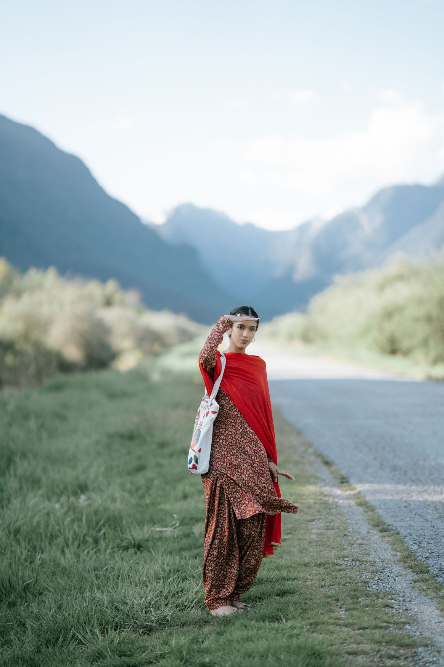 A person in a floral dress and red shawl walks along a mountain path surrounded by misty peaks and green grass.