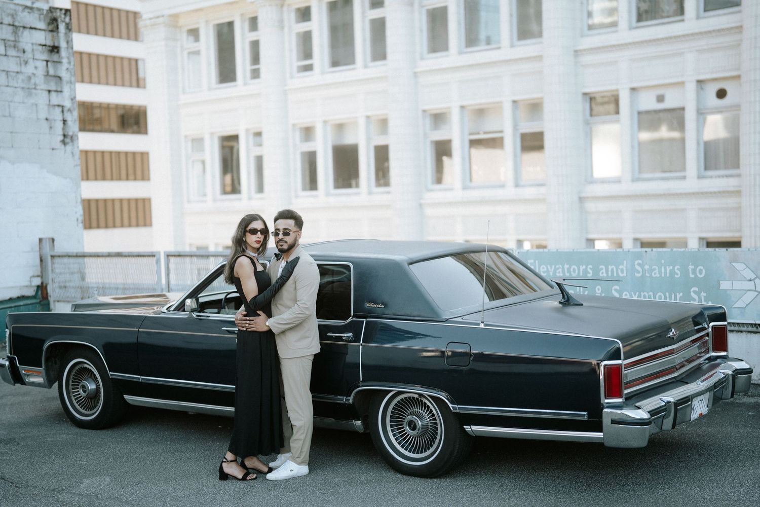 Classic black vintage car parked in front of white brick building with couple posing in elegant attire.