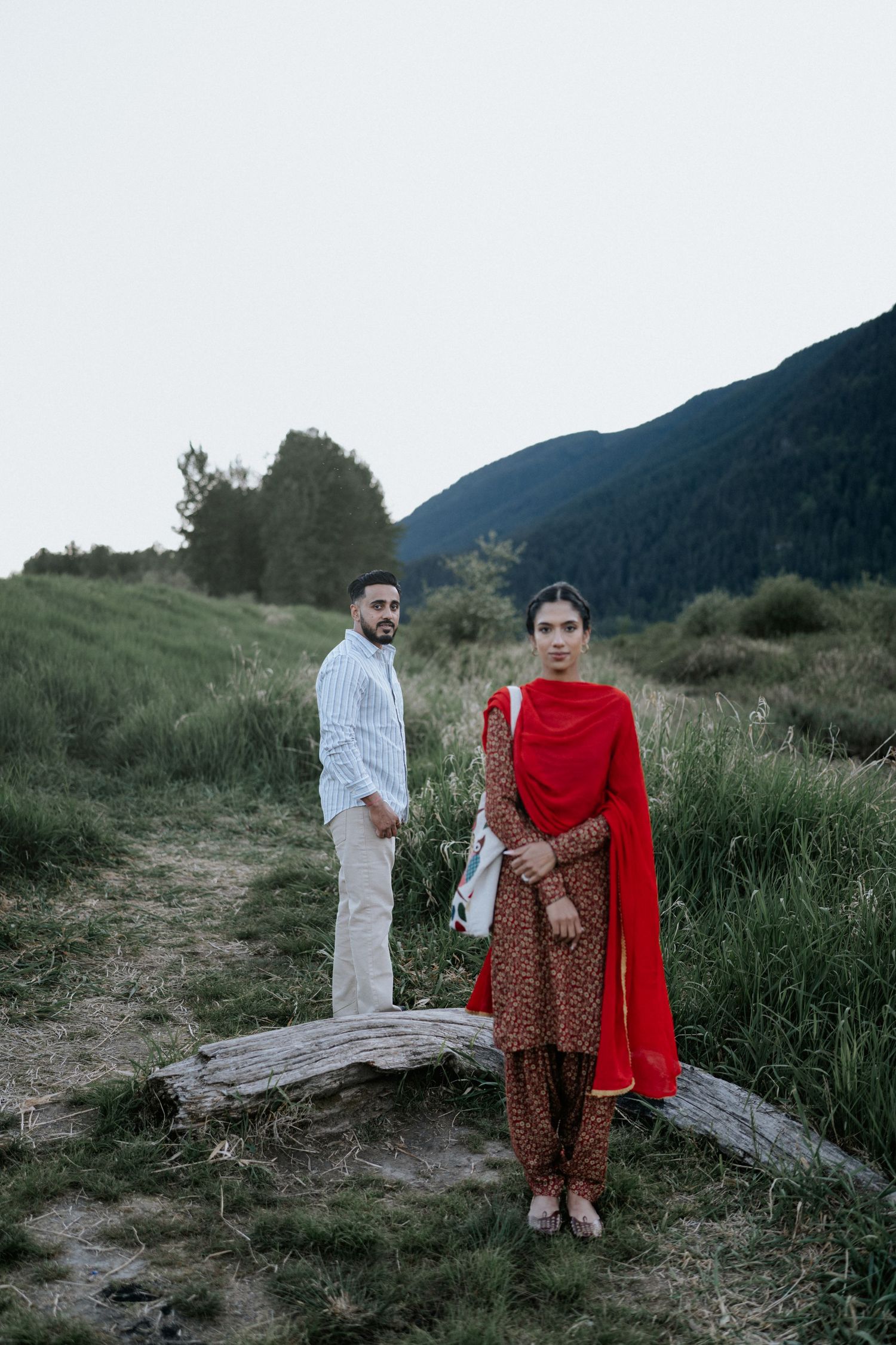 A couple stands together in a natural outdoor setting with mountains and tall grass in the background.