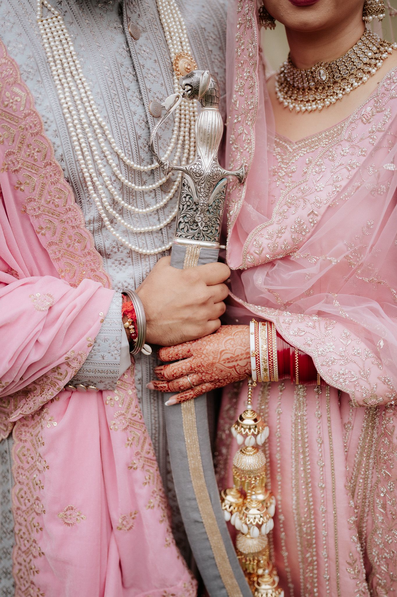 Close-up detail of traditional Indian bridal attire showing pink lehenga, pearl necklaces, mehndi hands and ornate jewelry.