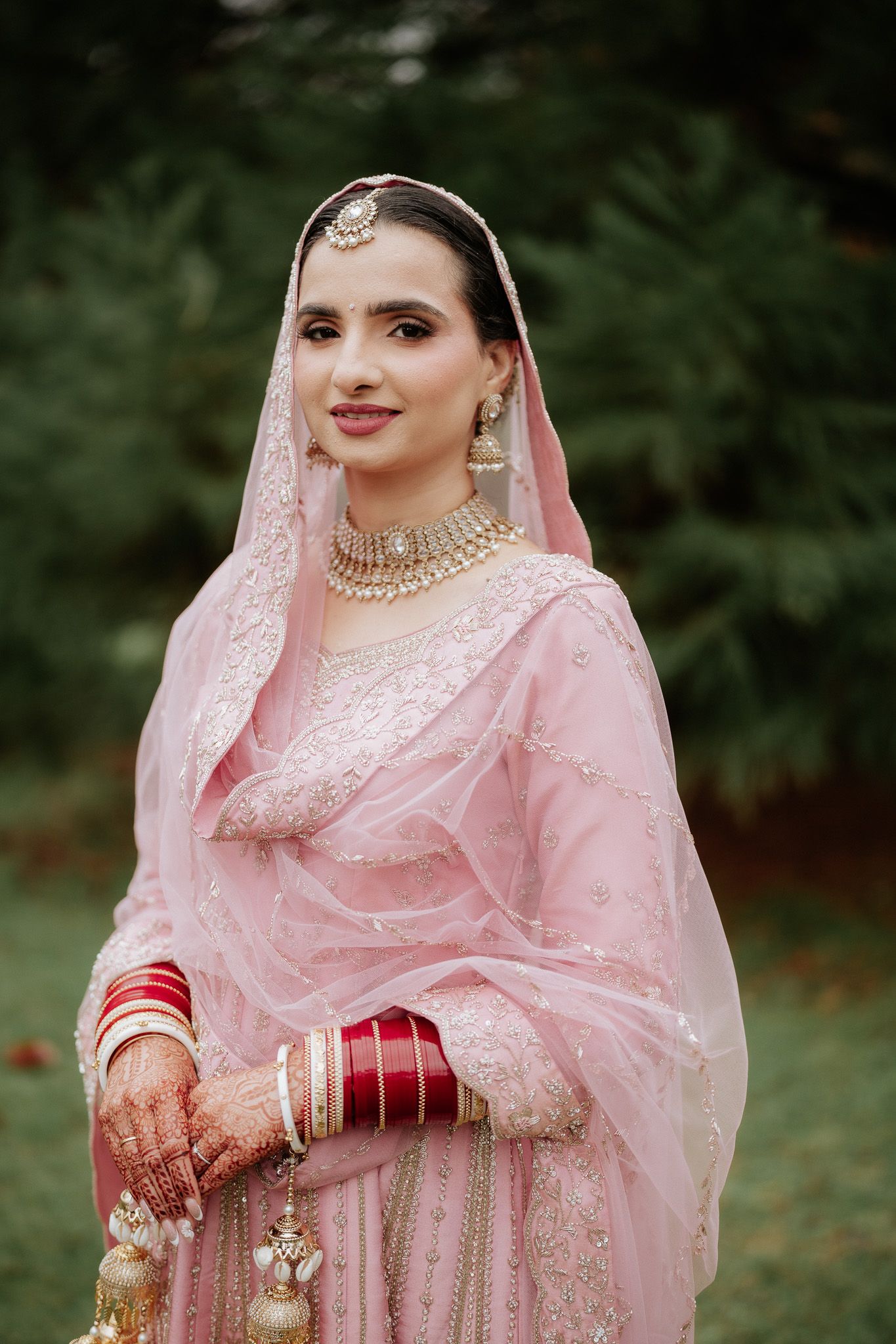 Traditional Indian bride in pink embroidered lehenga with sheer dupatta and ornate jewelry poses in garden setting.