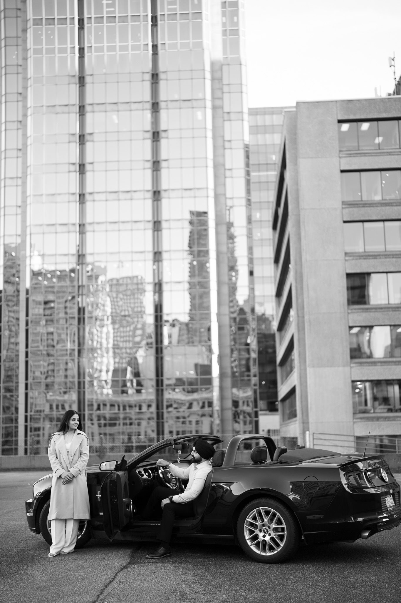 Black and white urban photo showing a luxury car parked next to a modern glass building in a city setting.