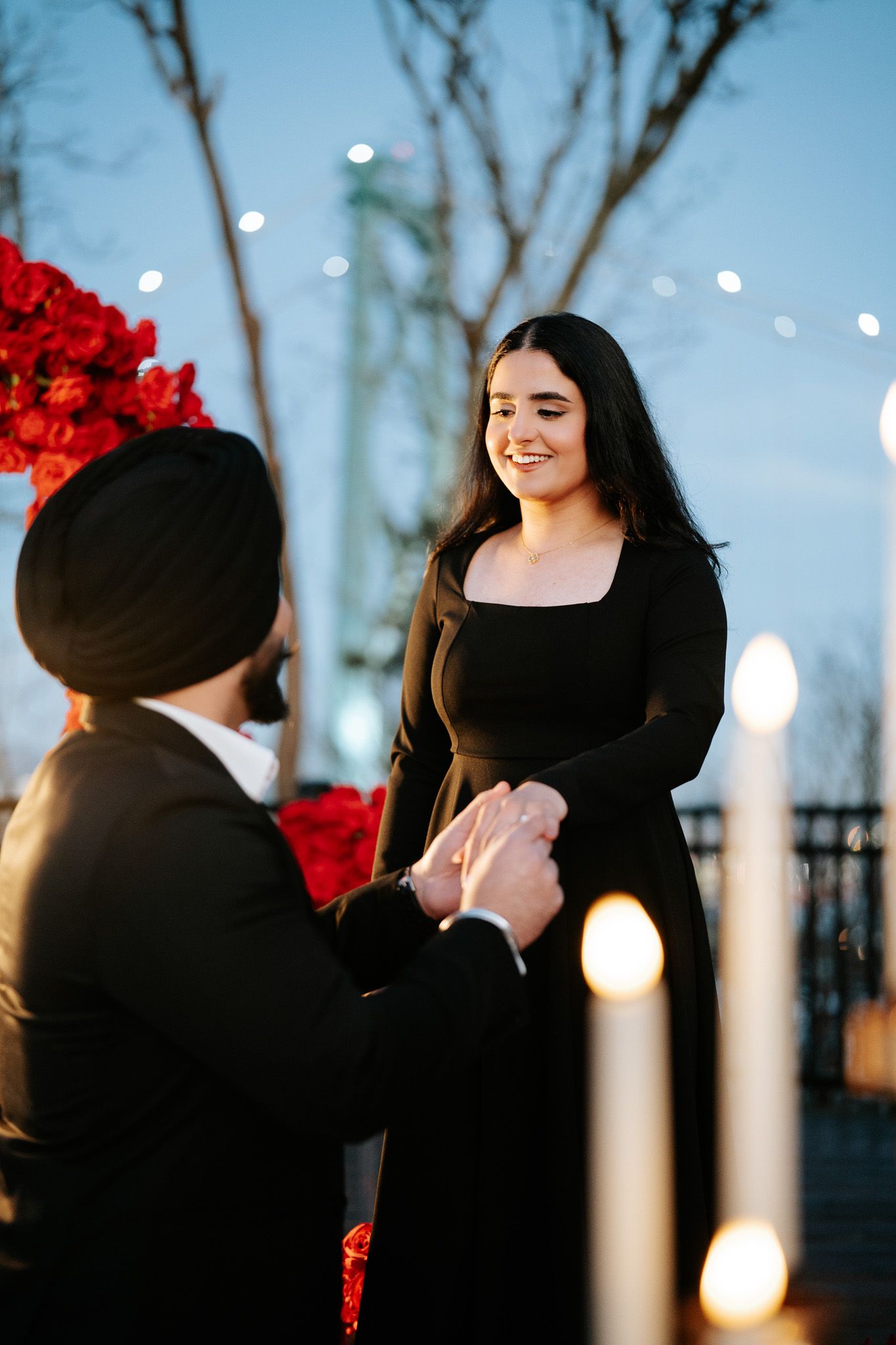 A romantic outdoor proposal scene at dusk with twinkling lights and red floral decorations in the background.