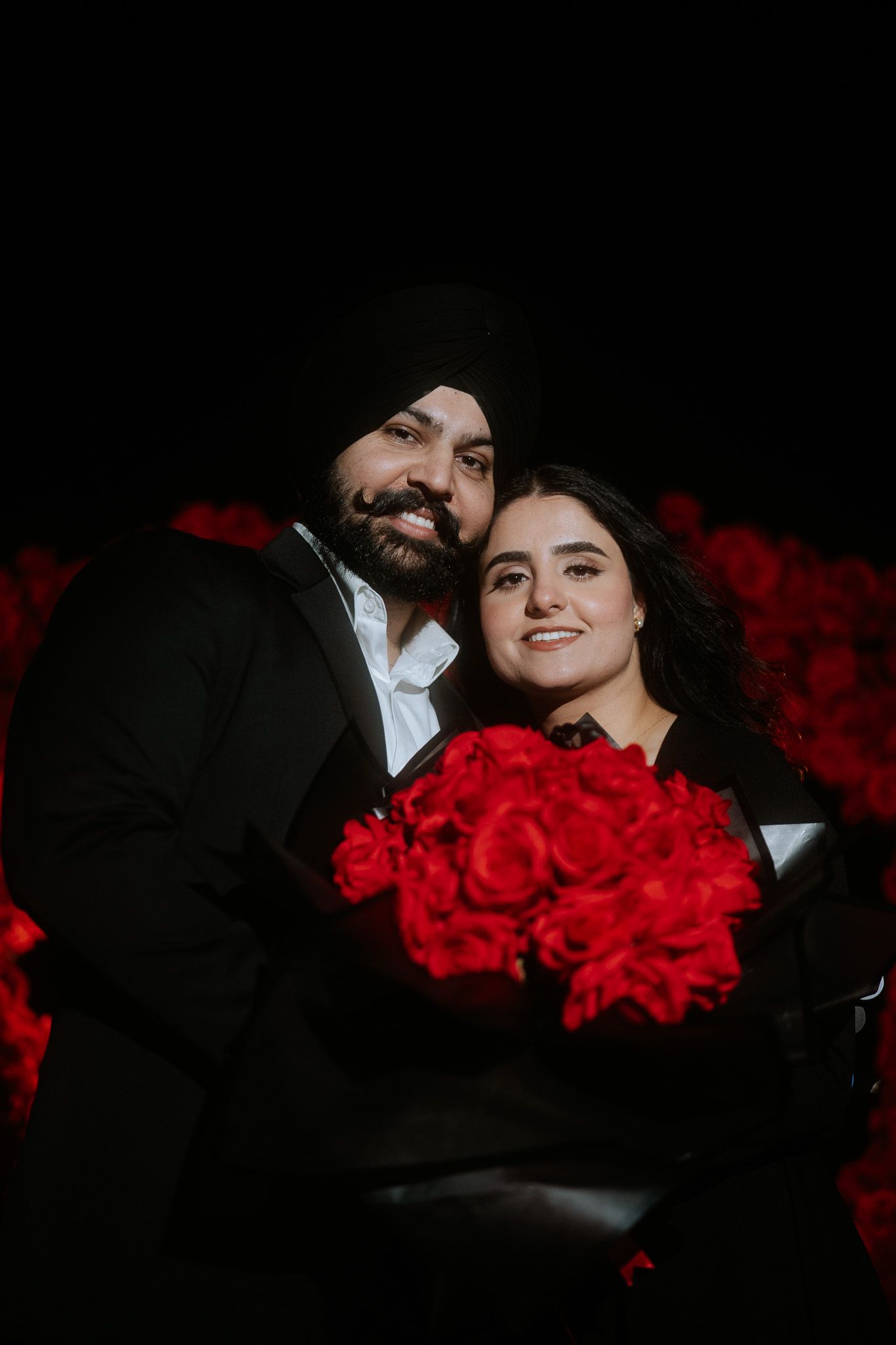 Romantic couple poses with vibrant red roses in dark setting creating dramatic contrast between light and shadow.