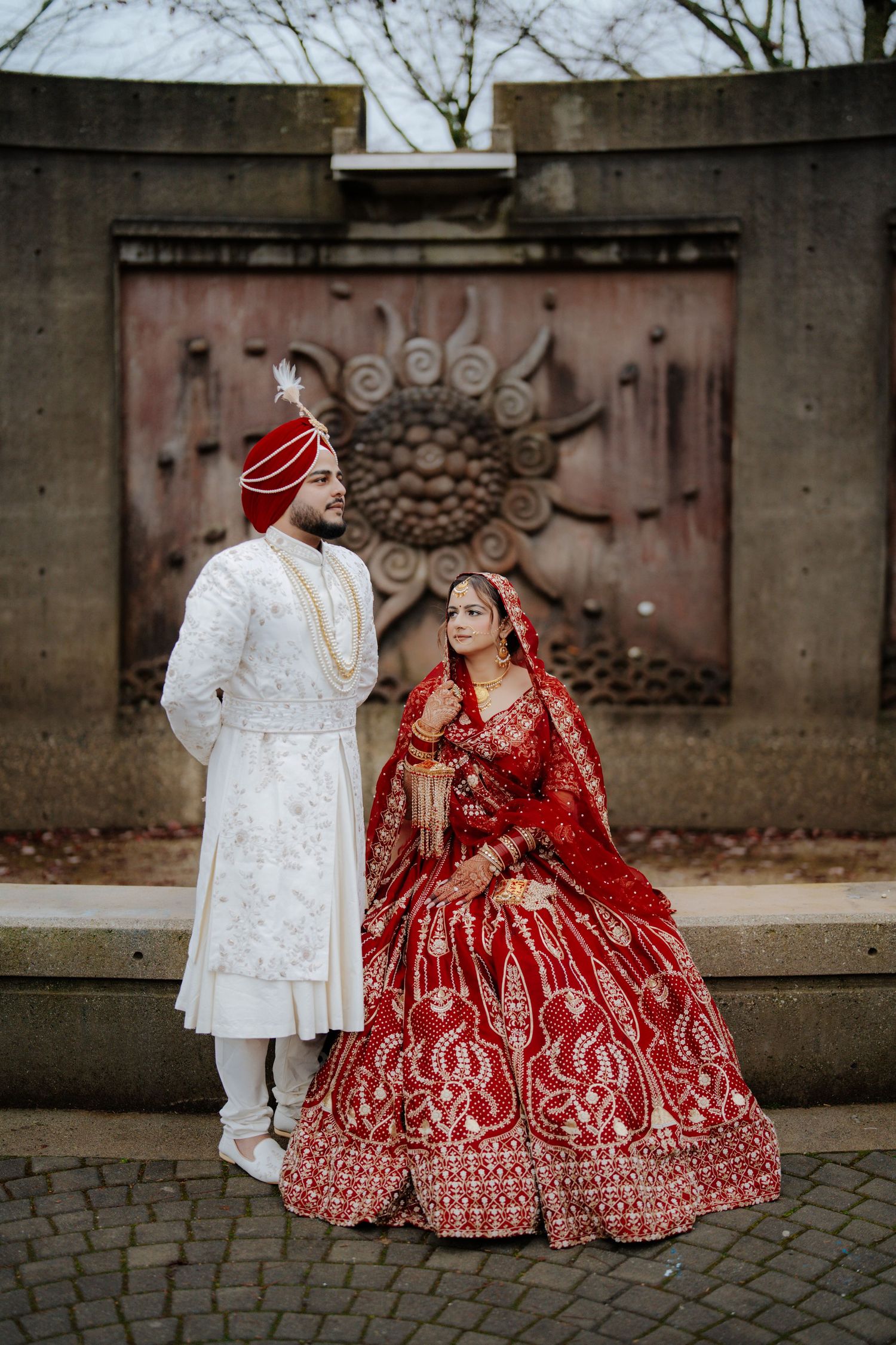 Traditional Indian wedding couple in red and white outfits posing against stone architecture.