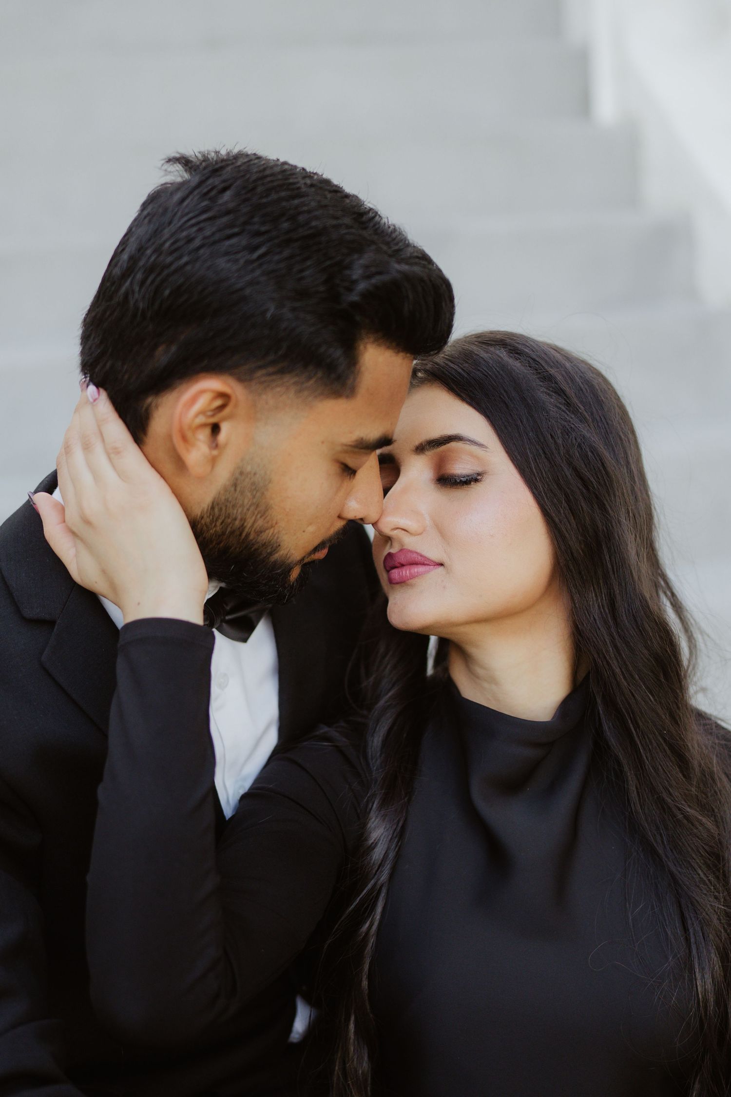 A romantic couple in black outfits share an intimate moment, touching foreheads tenderly on outdoor steps.