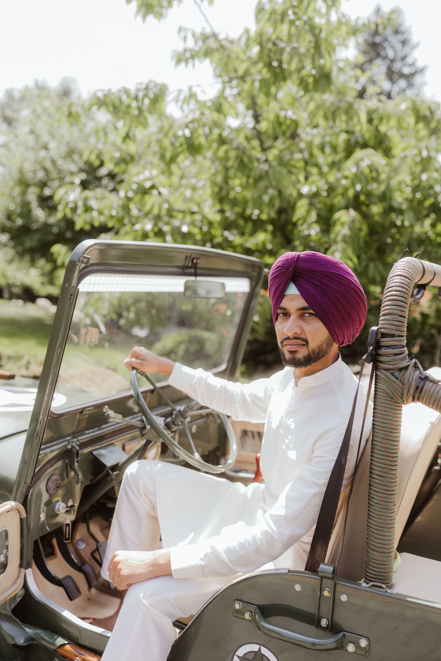 Sikh man in purple turban and white kurta sitting in a vintage military jeep outdoors.