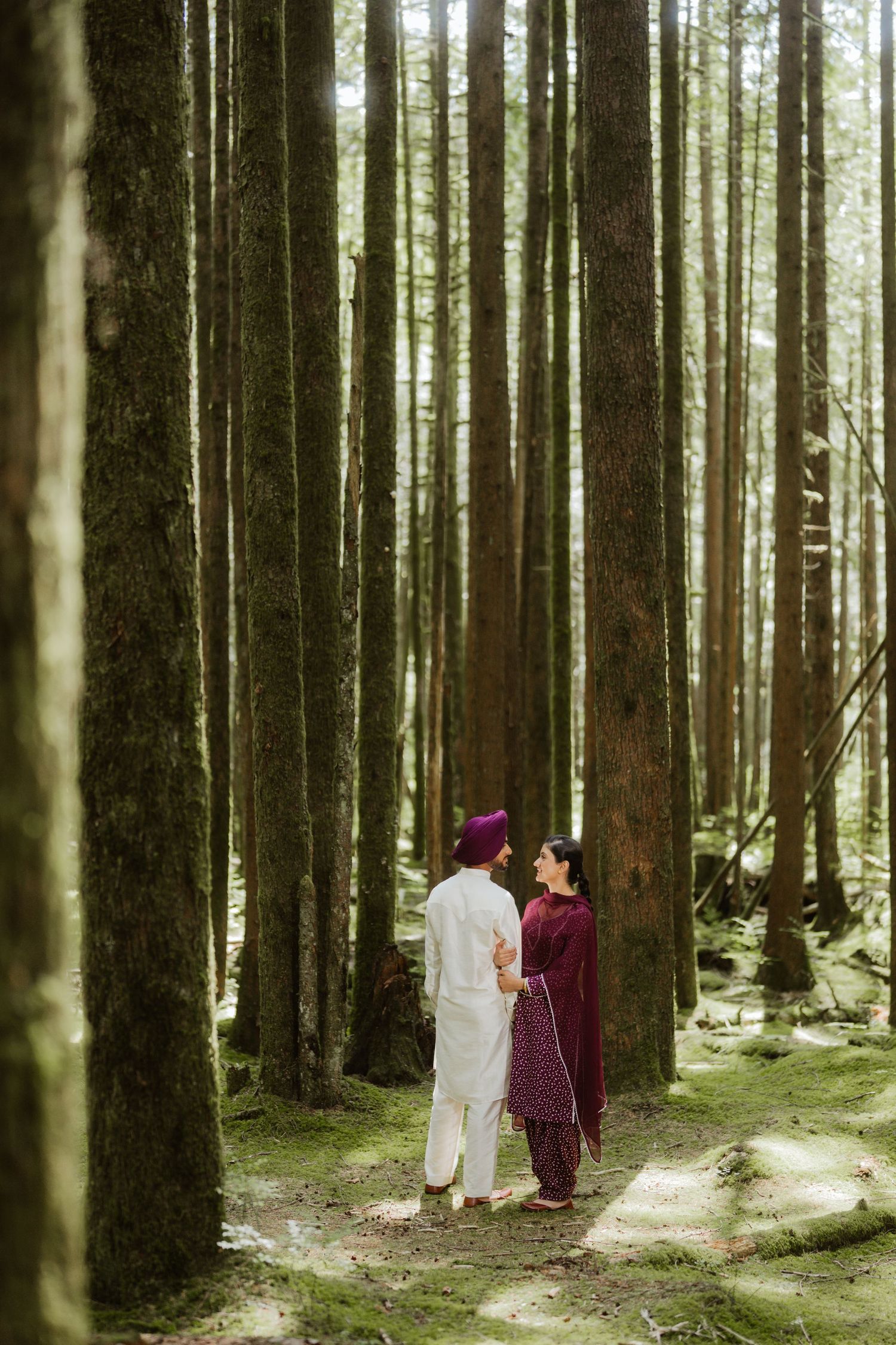 Sikh couple in traditional attire standing among tall trees in a lush forest during an engagement photoshoot.