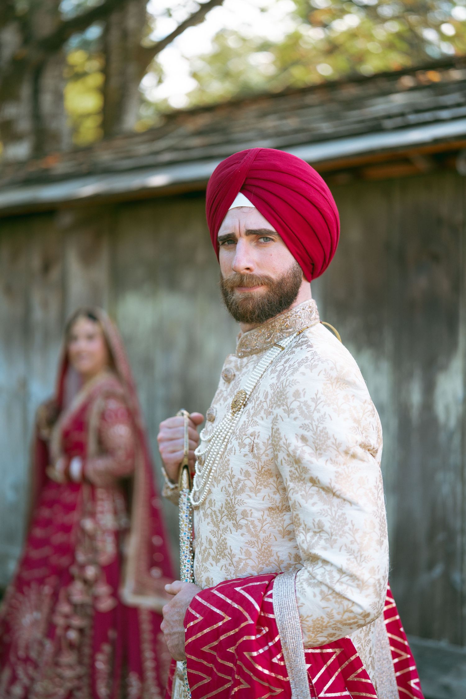 Sikh groom in red turban and gold sherwani stands before bride in red lehenga at outdoor wedding.