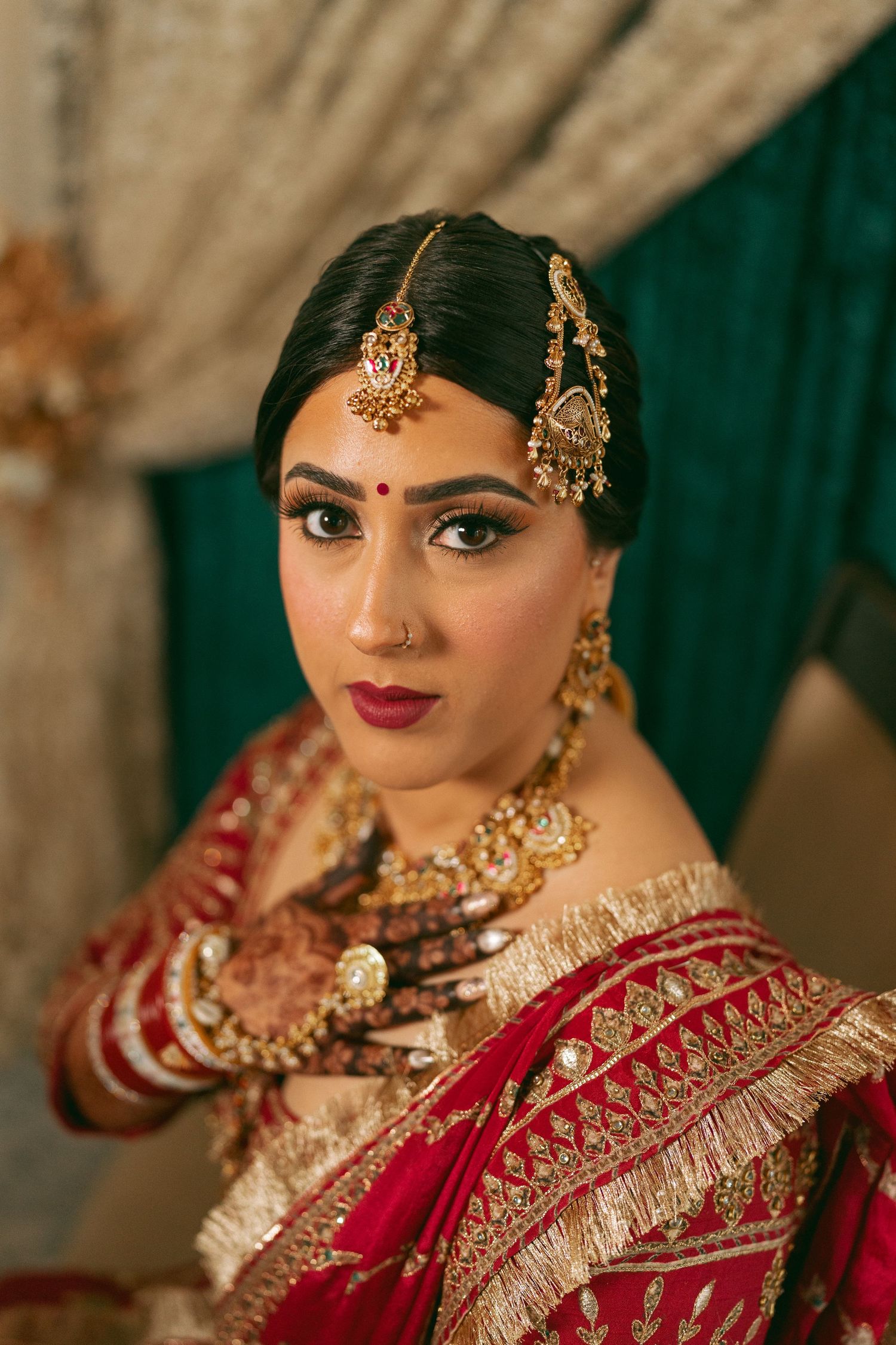 Indian bride in red and gold saree with traditional jewelry, maang tikka, and mehndi hands.
