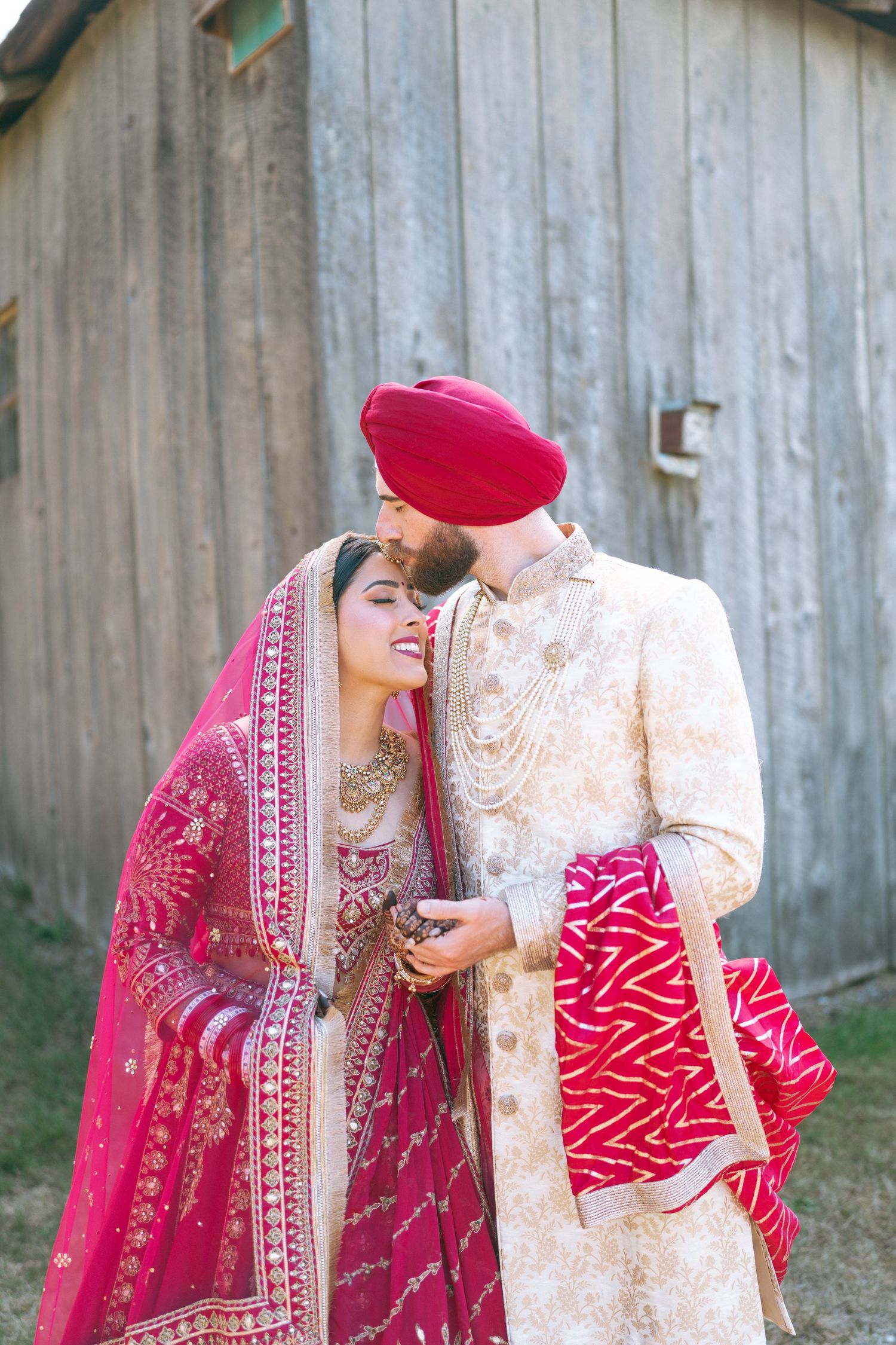 Sikh bride in red lehenga and groom in cream sherwani with red turban share a romantic moment at their wedding.