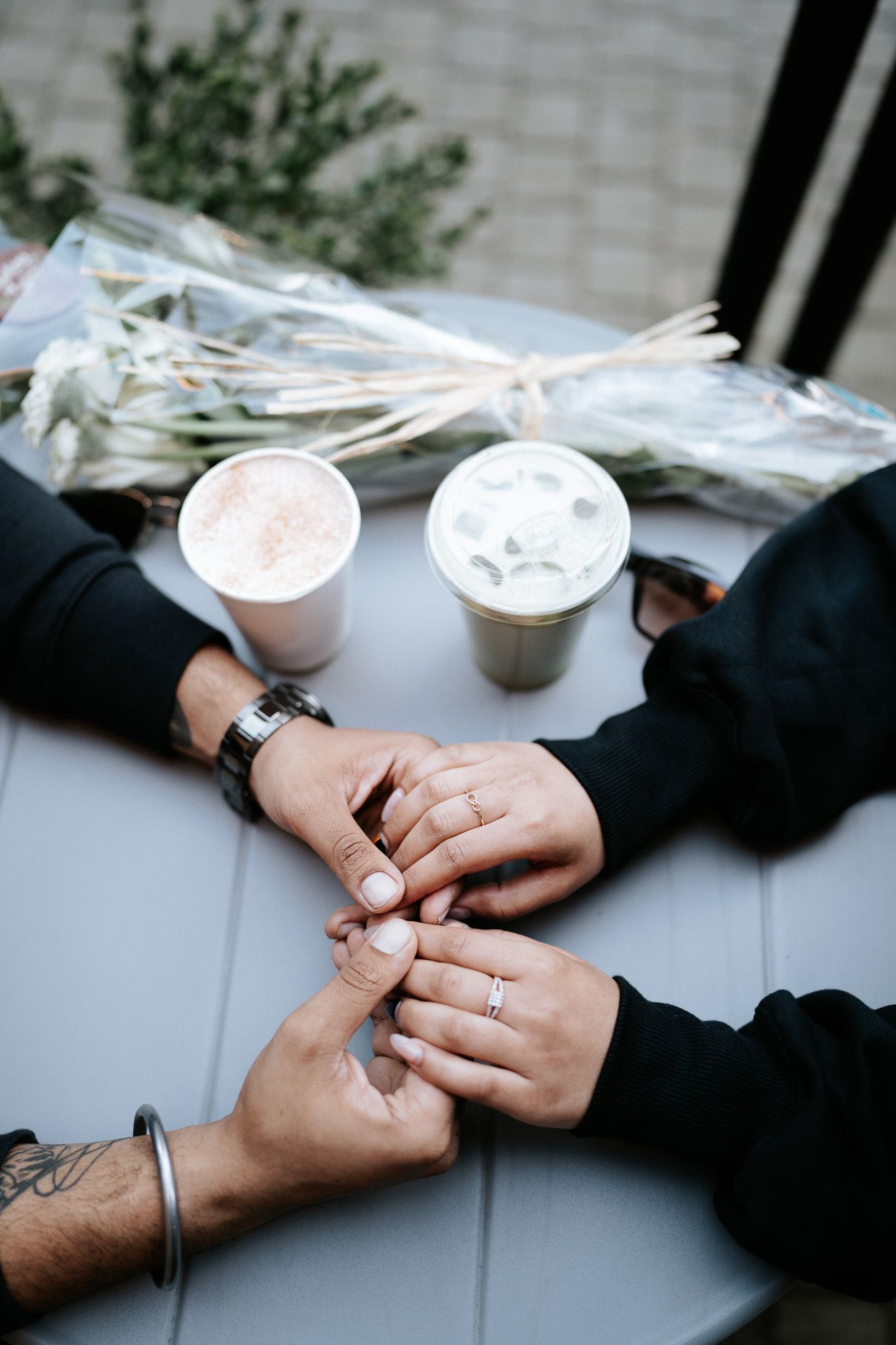Two people holding hands with engagement rings over a table with coffee drinks and flowers in the background.
