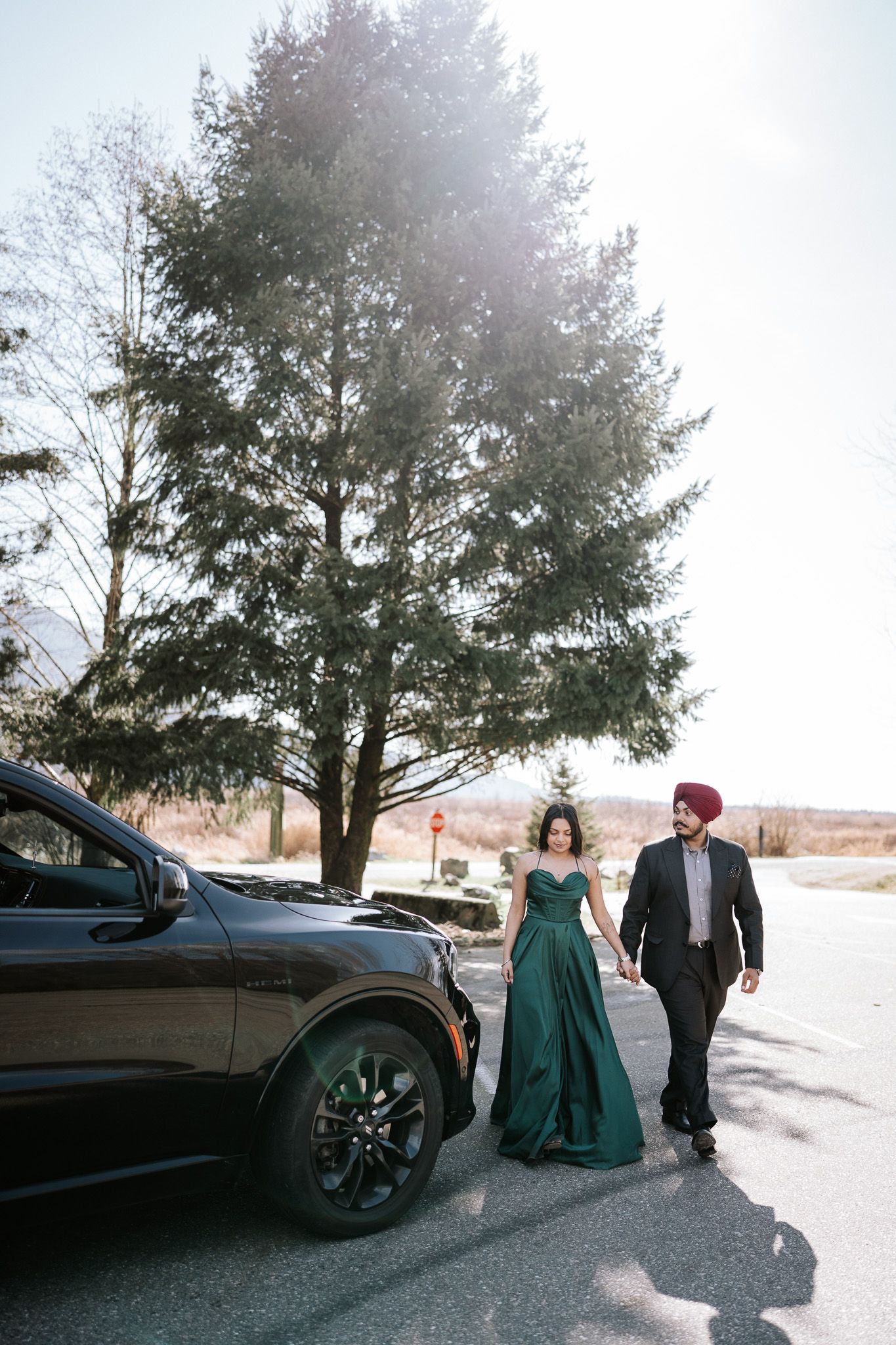 A couple in formal winter attire poses beside a black car in a snowy landscape at sunset.
