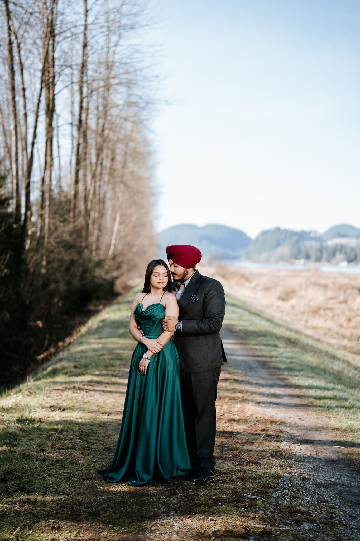 A couple shares an intimate moment on a tree-lined path with mountains visible in the distance.