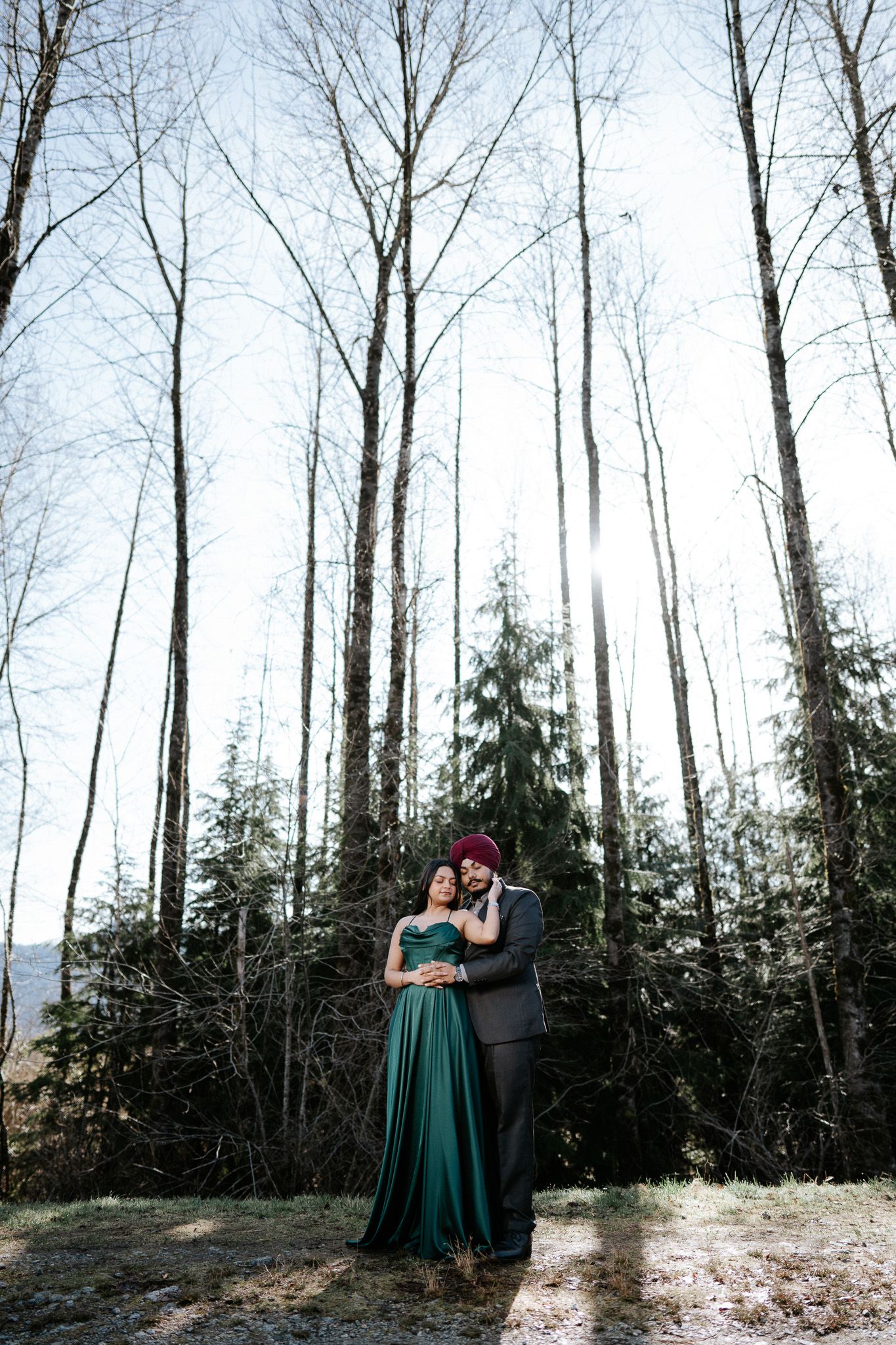 Couple shares intimate moments in a winter forest setting with bare trees and evergreens in the background.