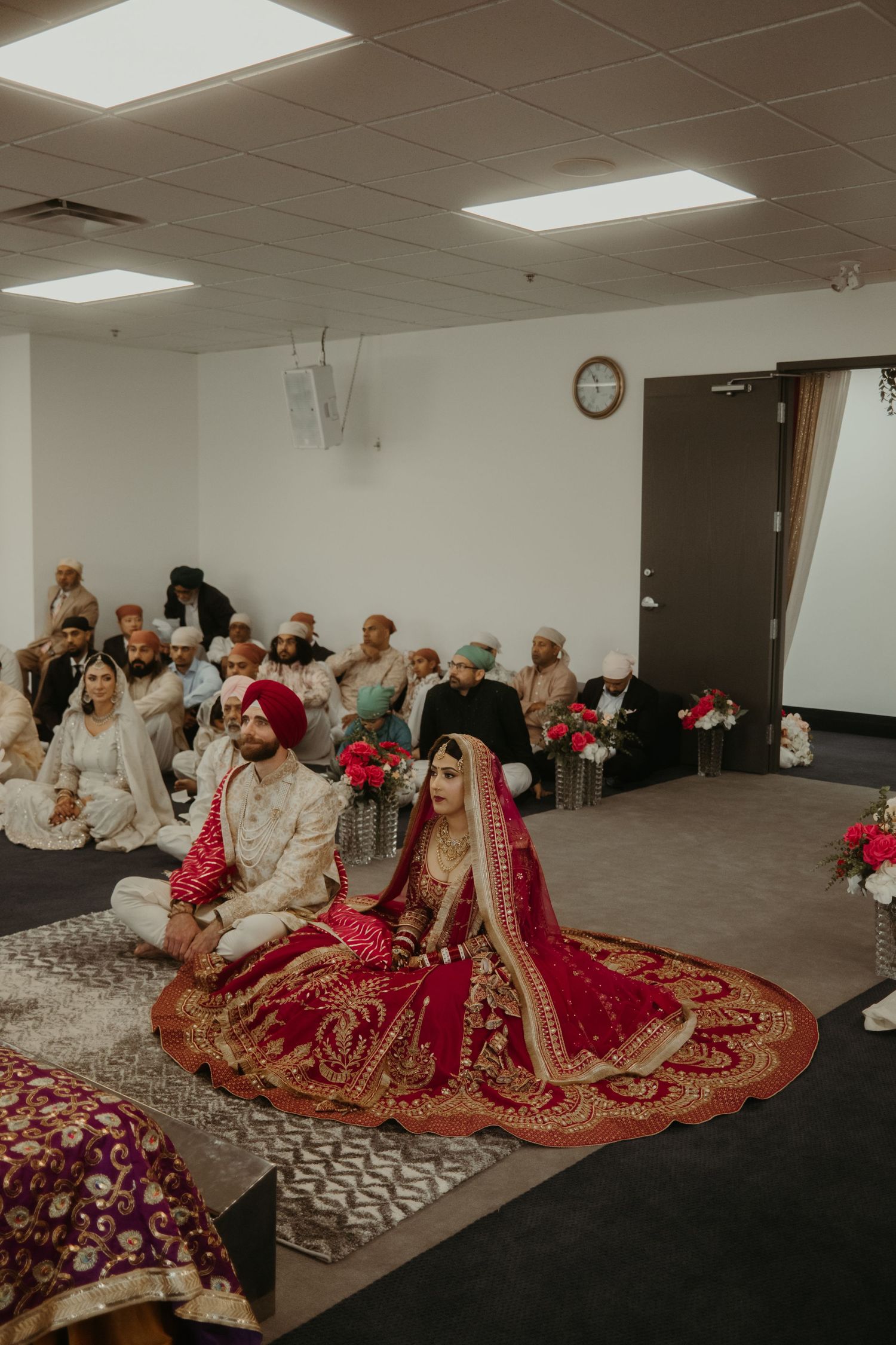 Sikh bride and groom in red and gold attire sit during Anand Karaj wedding ceremony with guests seated behind them.